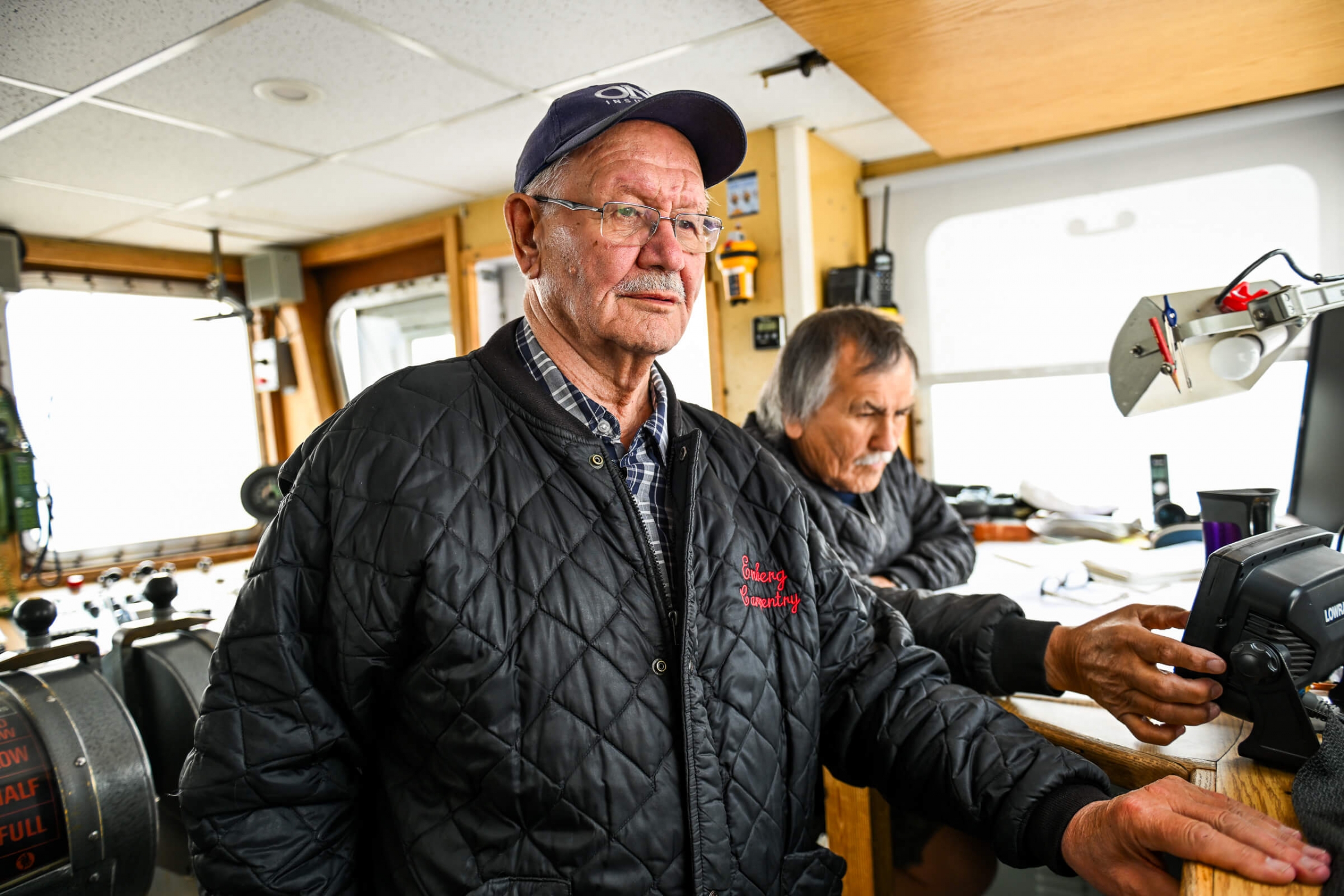 Two older men inside the cockpit of a boat, looking intently at the instruments to help the navigate