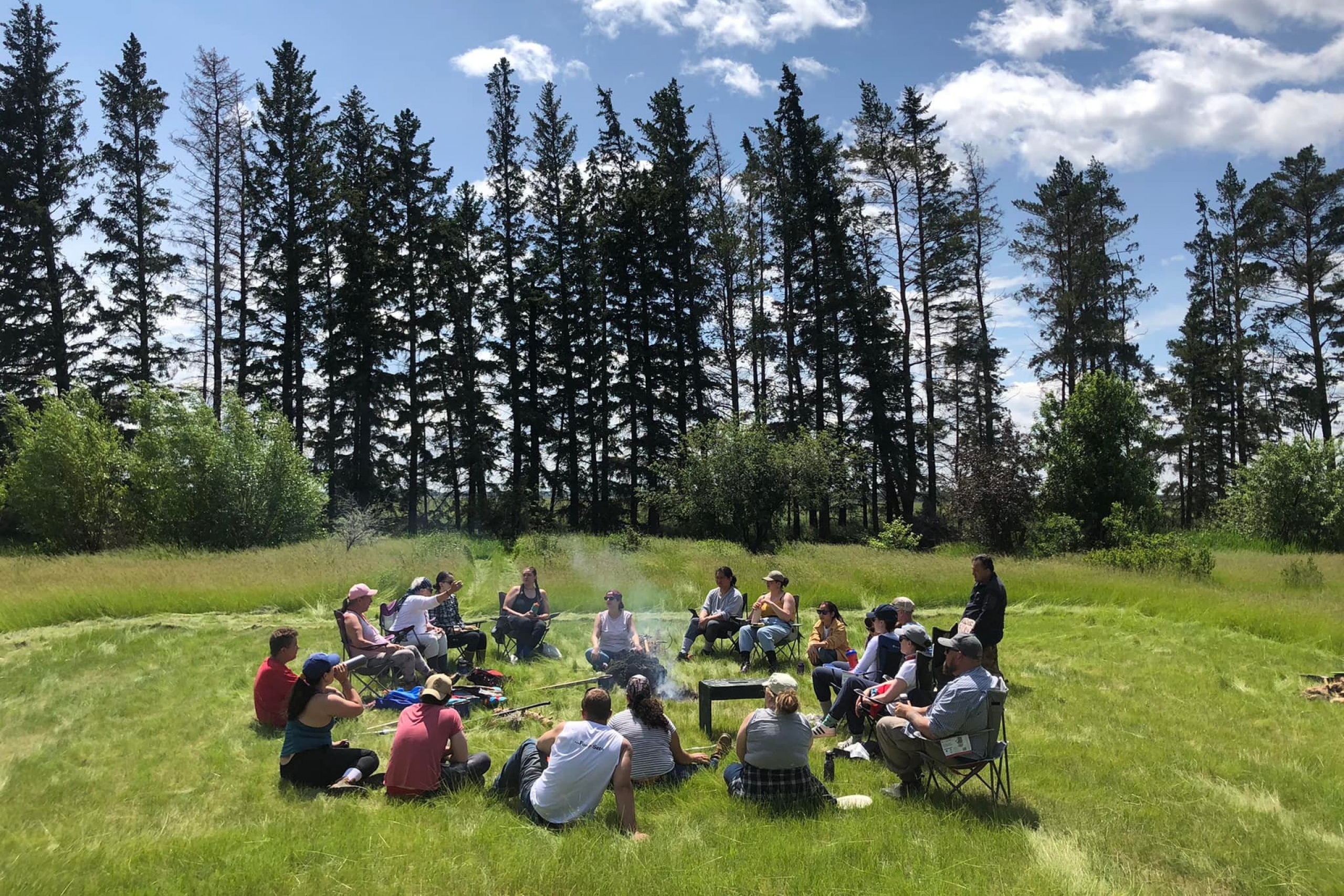 group sitting in a circle in a field around a fire on a summer day.