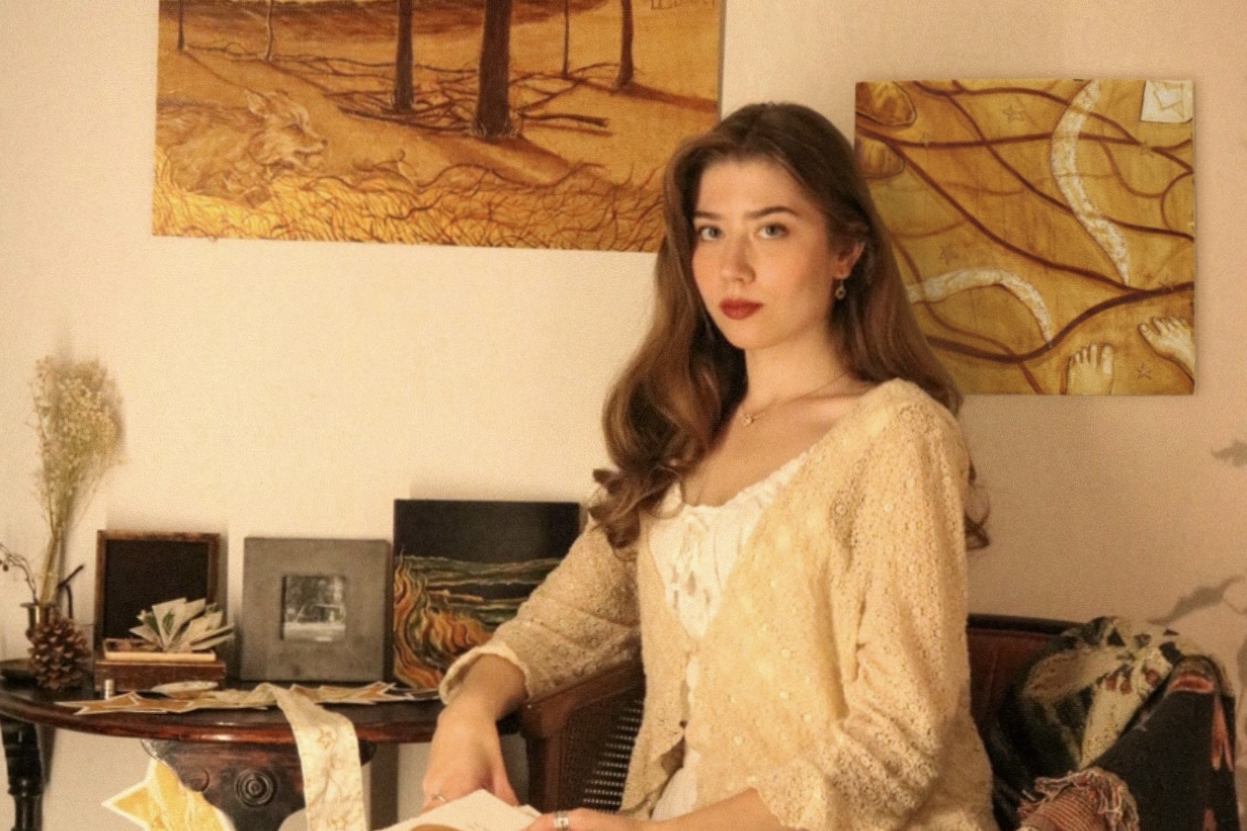 Leah Gertzen sits in a warmly lit studio, holding a small artwork, with her landscape paintings displayed on the wall behind her.