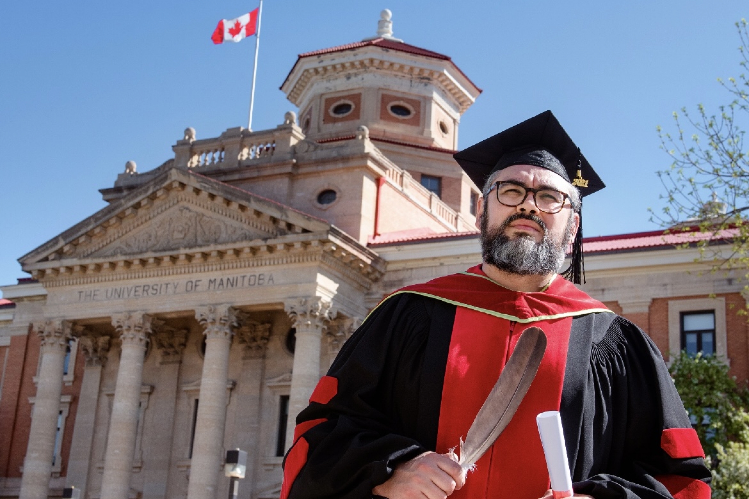 Man holding eagle feather and scroll, wearing a graduation cap and gown in front of UM building.