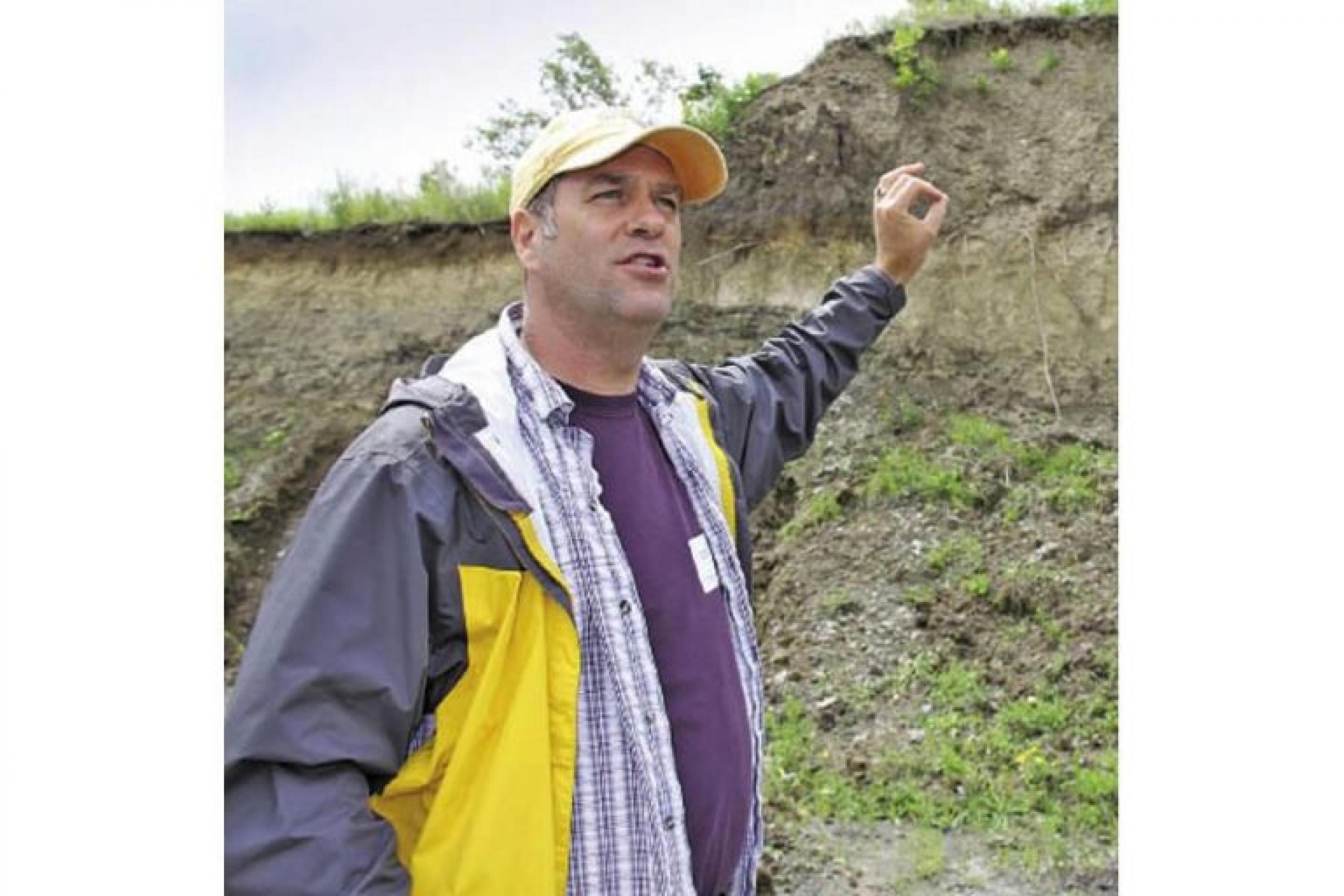 A man (Dr. David Lobb) in a yellow baseball cap and a raincoat stands in a valley