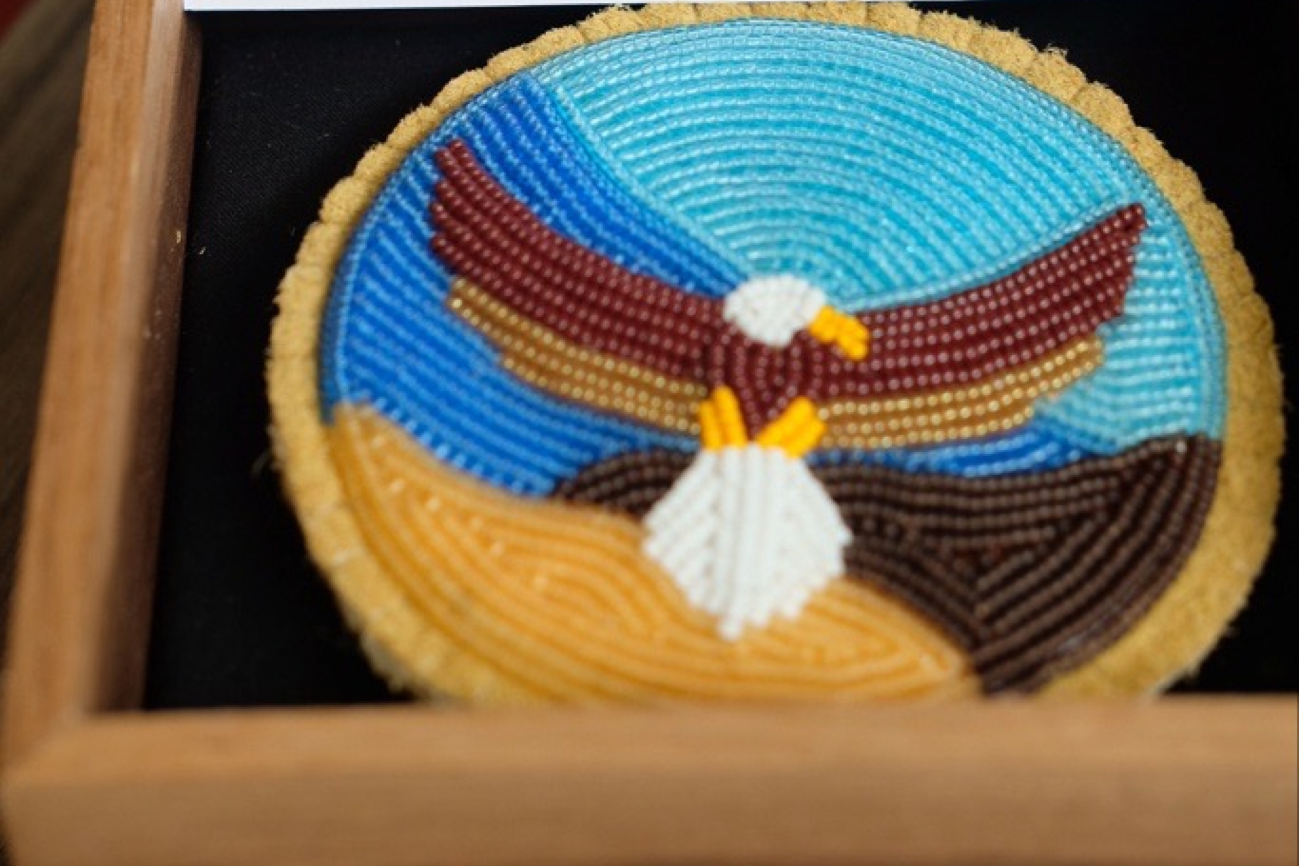 Beaded medallion of eagle against blue sky and brown and gold landscape, placed in small square wooden box.