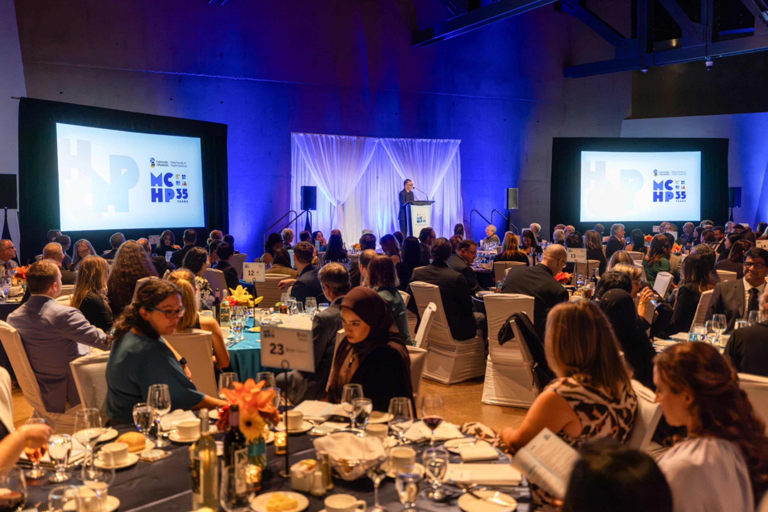 Gala dinner event with a speaker at a podium. Two large screens display the text: “University of Manitoba, Rady Faculty of Health Sciences, MCHP, 35 Years.”