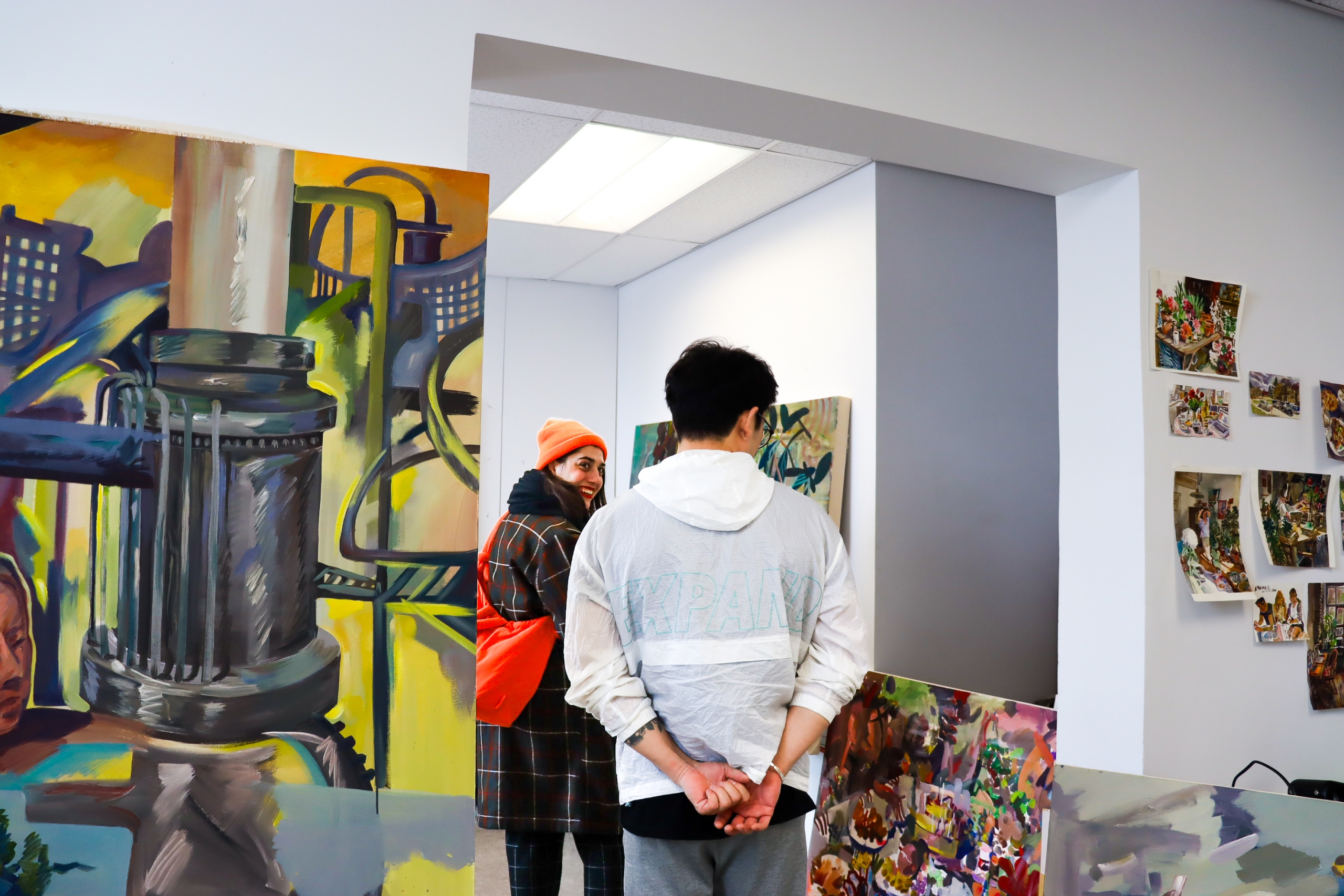 Two visitors stand in an MFA studio looking at colourful paintings on large canvases, including a large urban scene painting leaning against the wall.