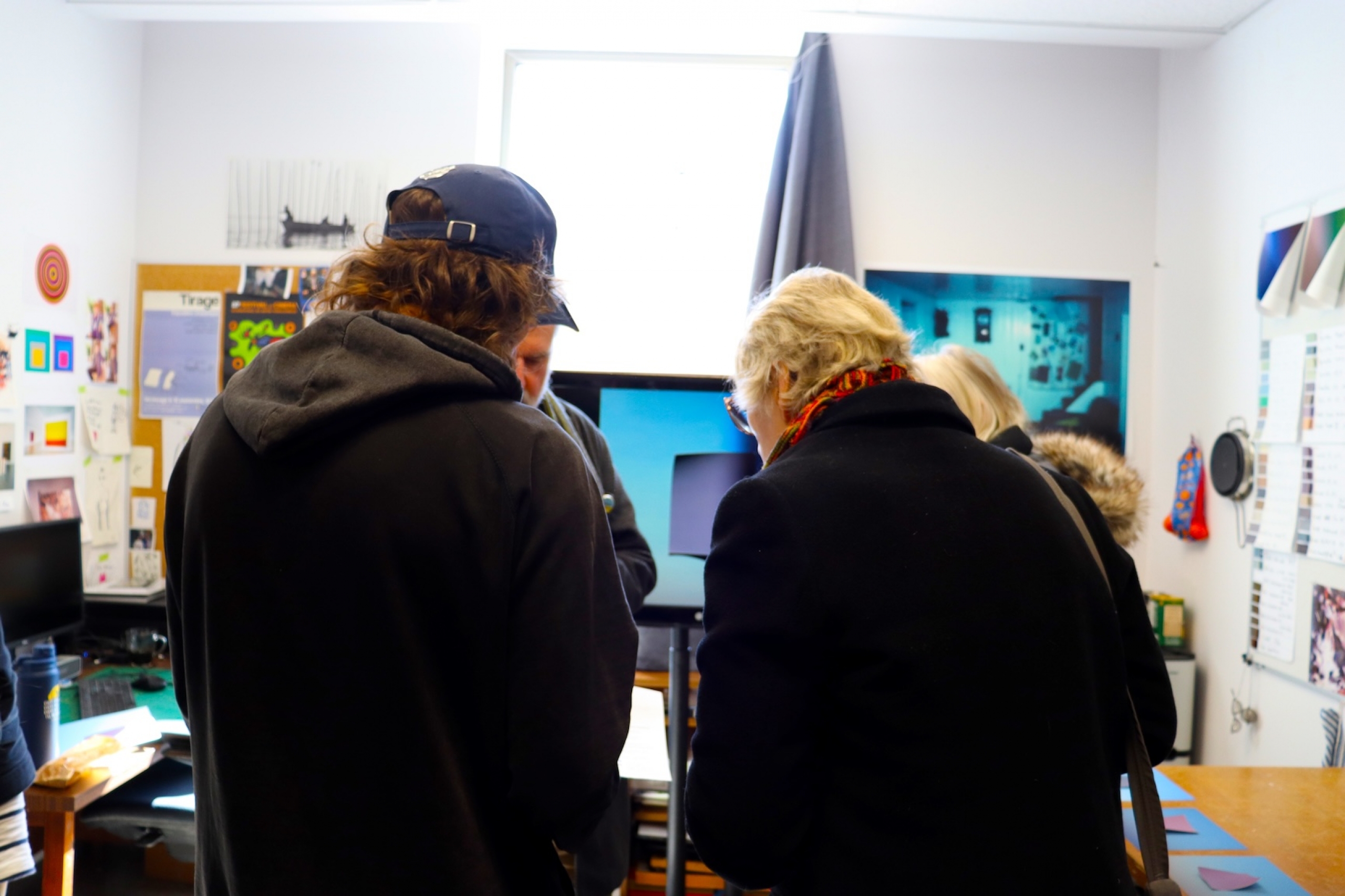 Two people stand in the doorway of an MFA studio discussing colourful paintings displayed on the walls, with additional artworks and materials spread across a table.