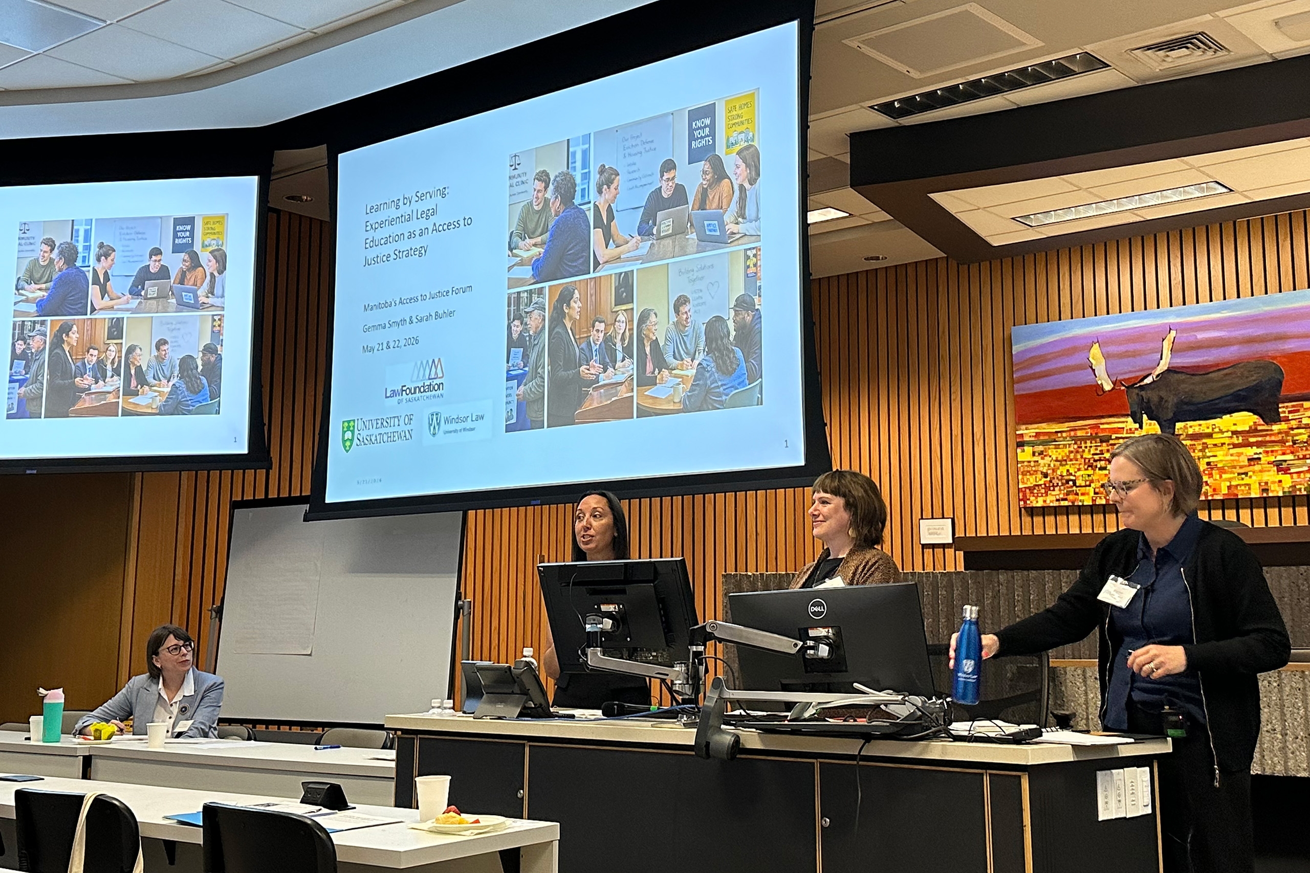 four women get ready to speak to a powerpoint presentation on a big screen behind them at the front of a large wood panelled room