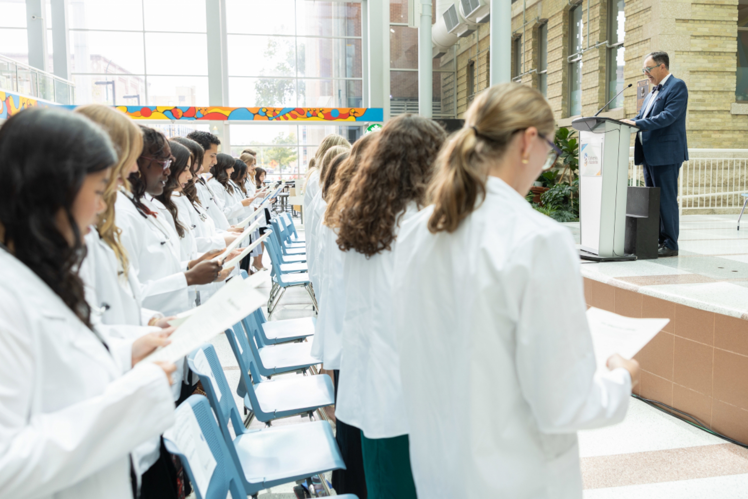 Dr. Peter Nickerson speaks at a lectern on a stage. The students are standing and holding papers.