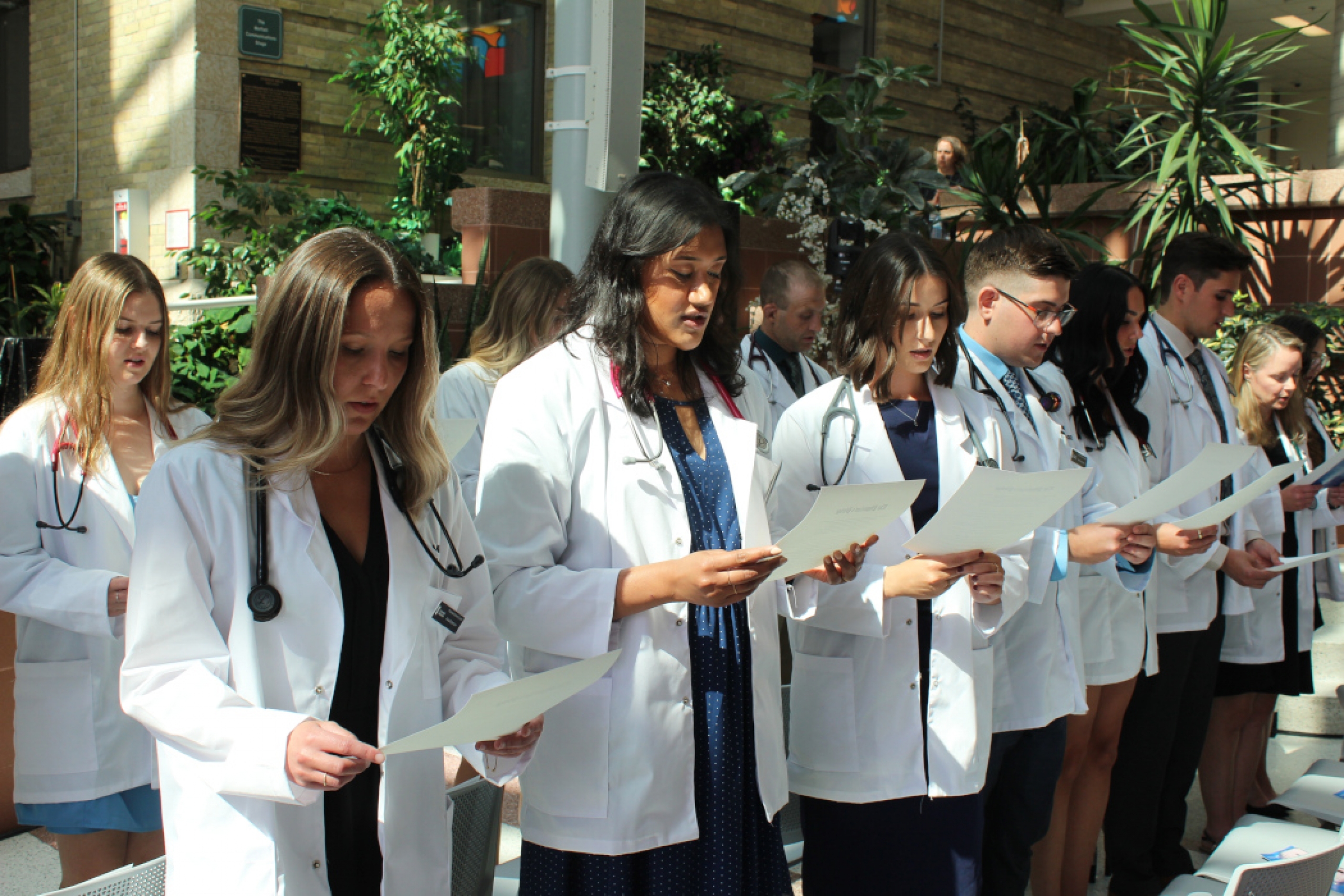 Two rows of students are wearing white coats and stethoscopes. They are standing and each is reading off a paper they are holding.