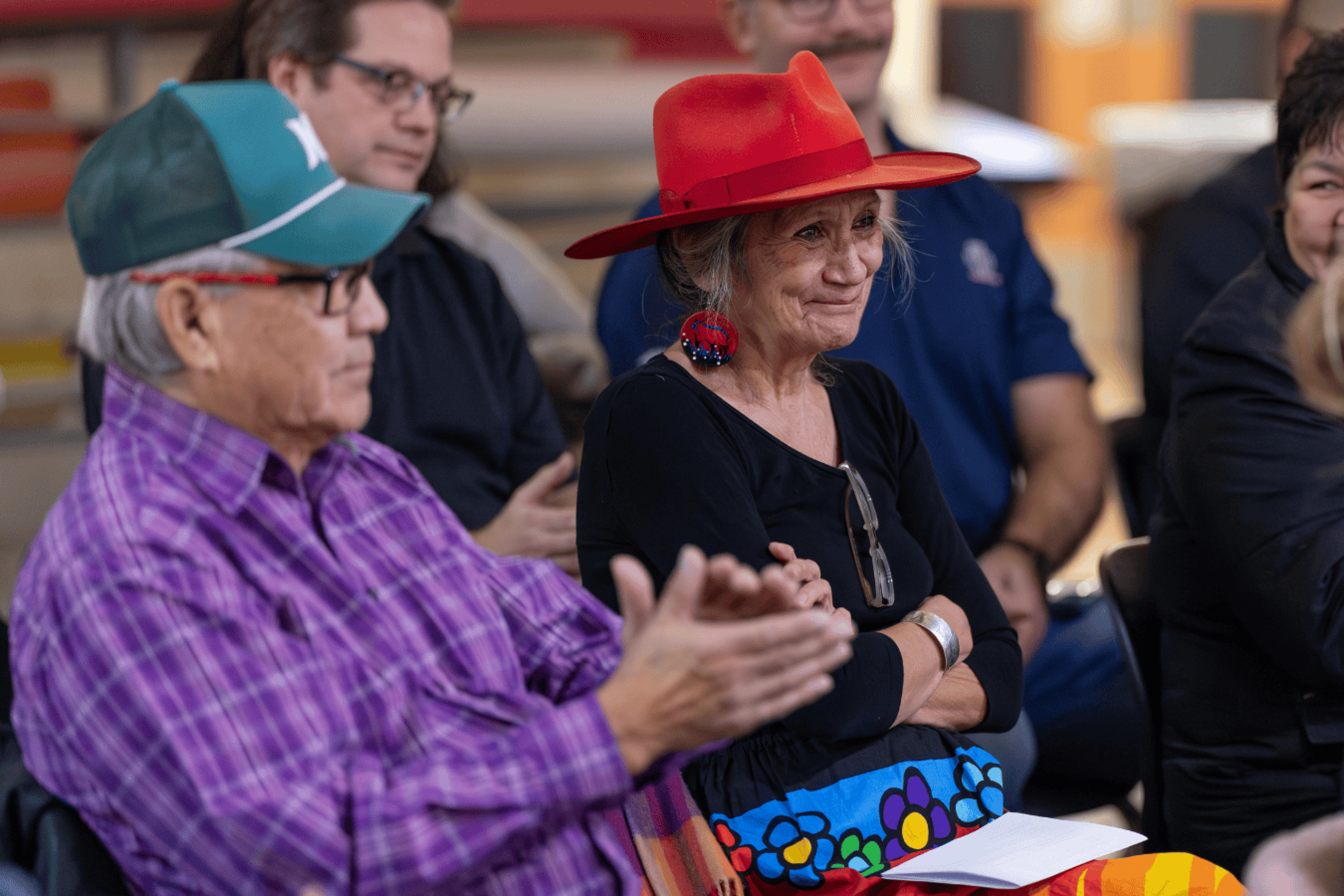Audience members seated and listening, including a woman in a red hat and beaded earrings.