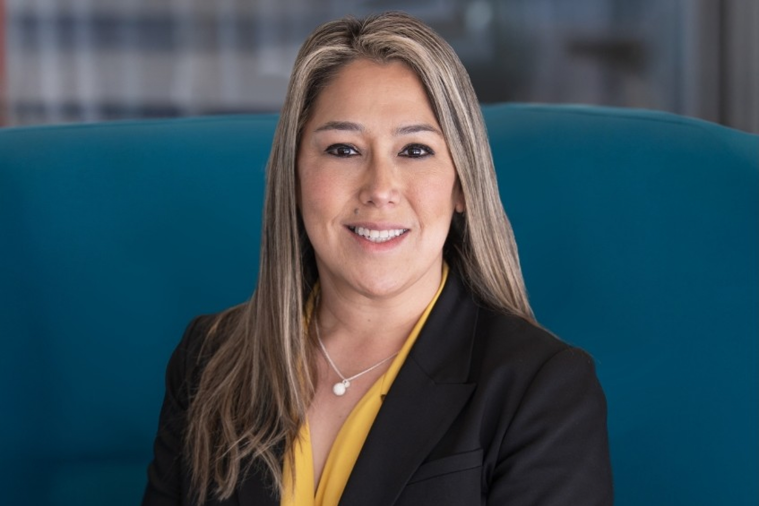 Woman in a black suit seated on a teal chair, smiling