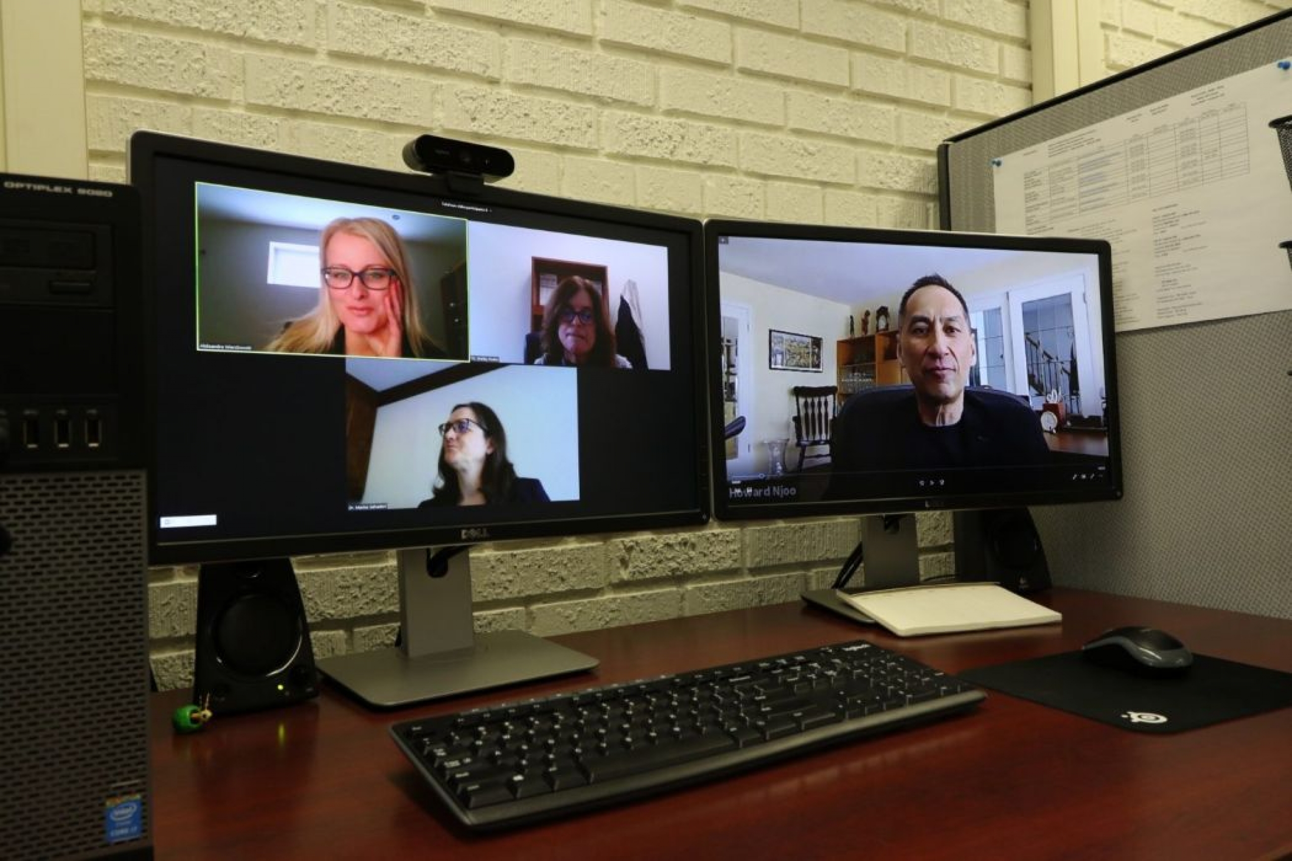 Two computer monitors with people on the screens. A keyboard and mouse sit on a desk. 