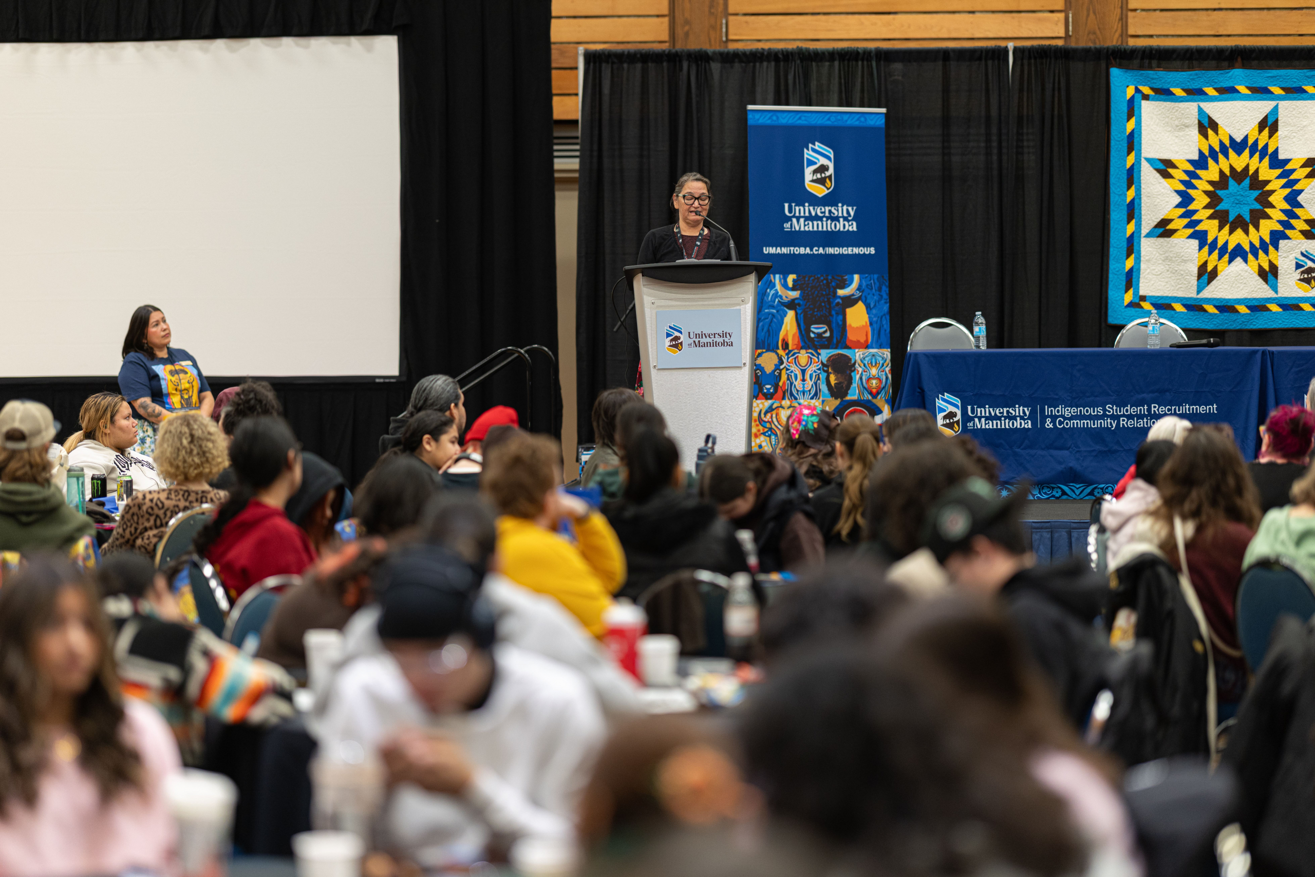 Kookum Karen stands on stage behind podium, addressing crowd of high school students.