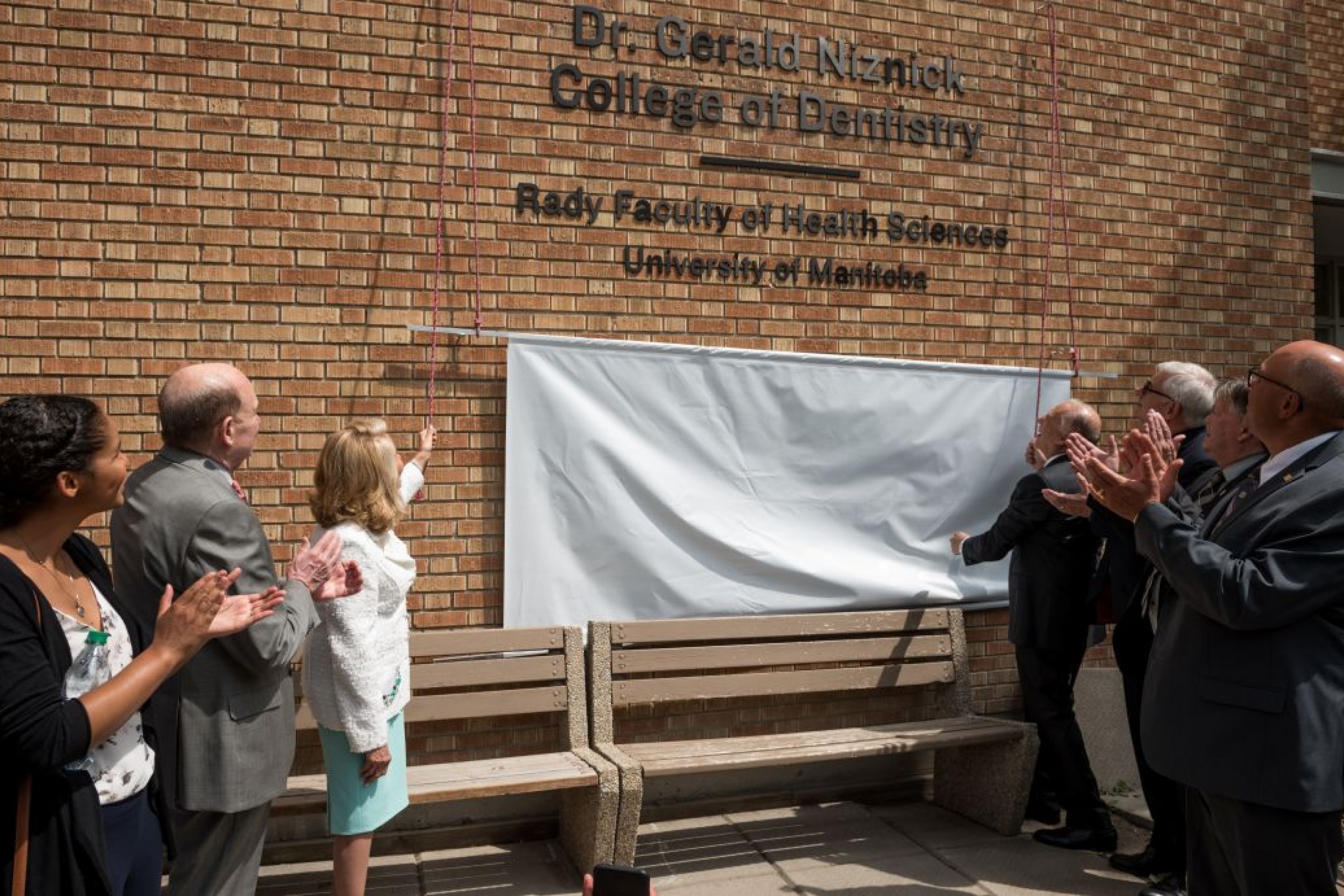 A sign on a wall is unveiled that reads "Dr. Gerald Niznick College of Dentistry. Rady Faculty of Health Sciences. University of Manitoba." People stand below the sign and clap. 