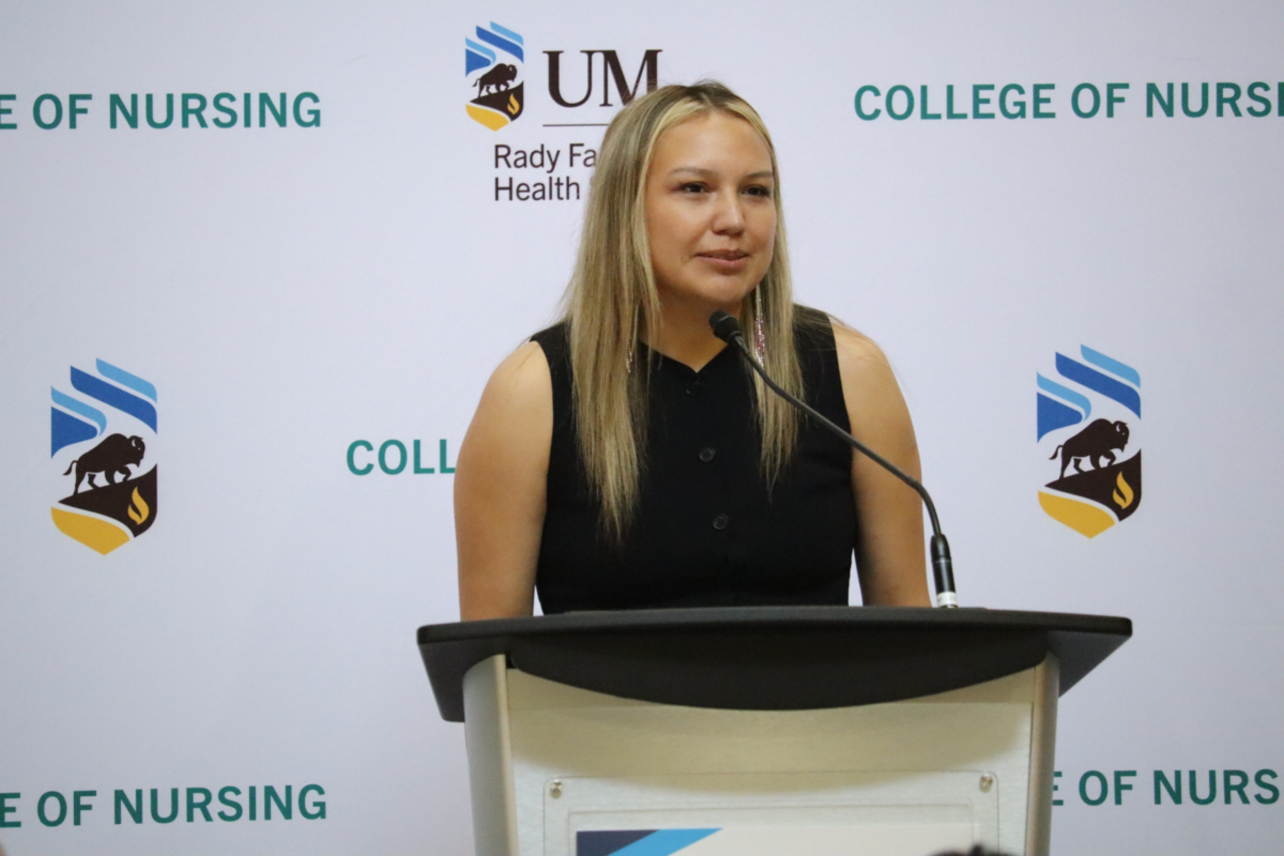 A person talks in front of a podium. Behind is a backdrop with UM Rady Faculty of Health Sciences logo and College of Nursing.