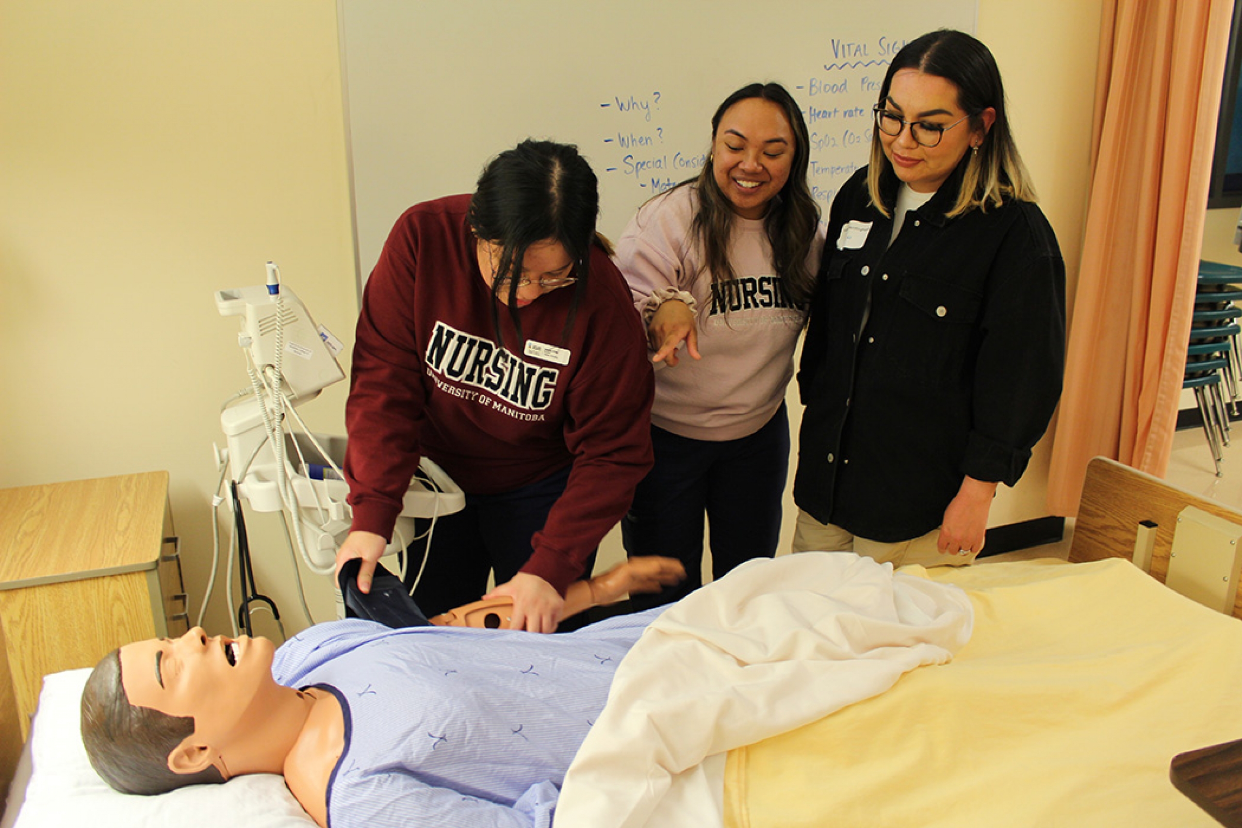 Three female nursing students practice skills in a simulated hospital room with a manikin.