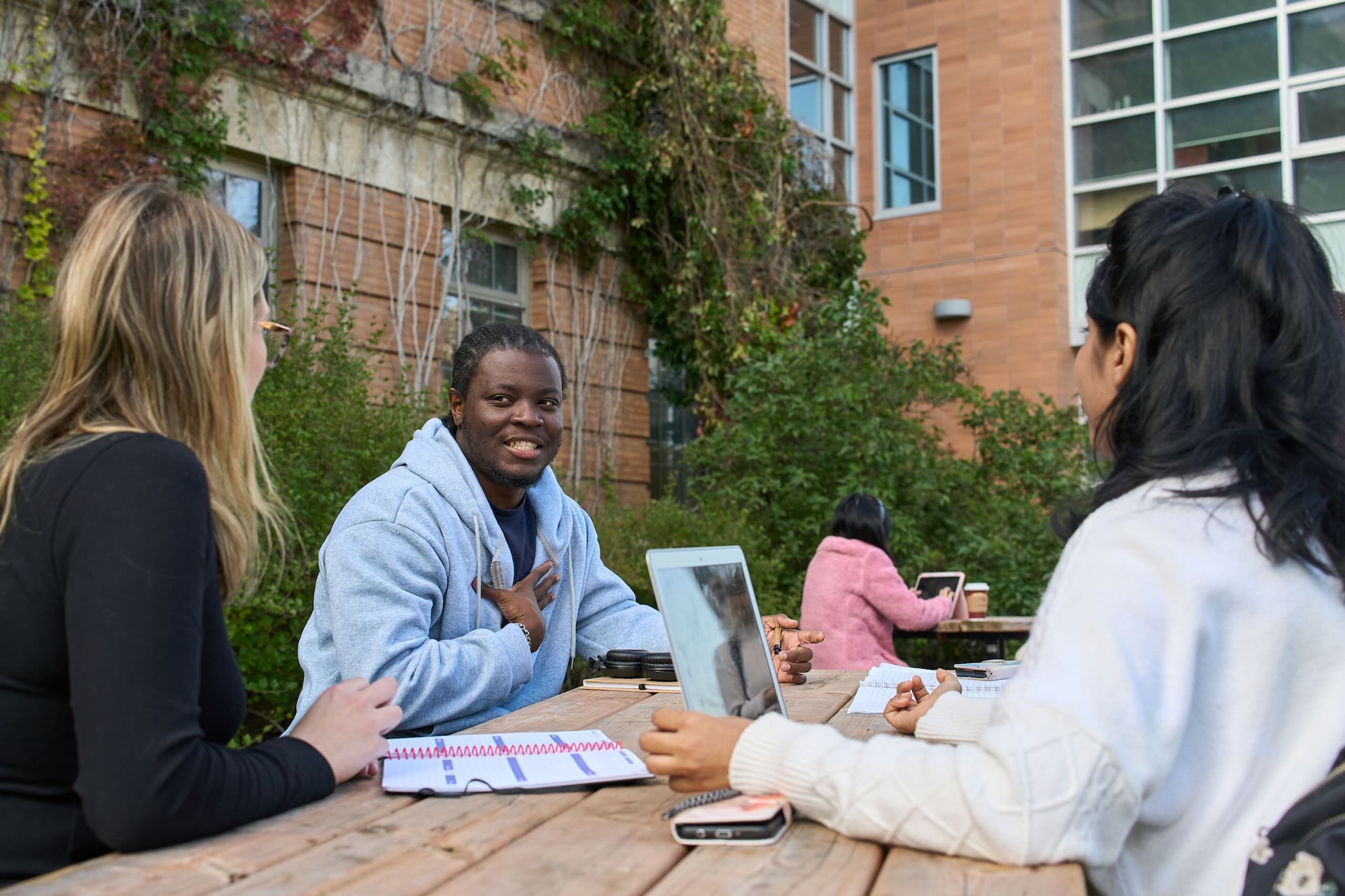 Black smiling male student sits at an outdoor picnic table with other students and a computer.