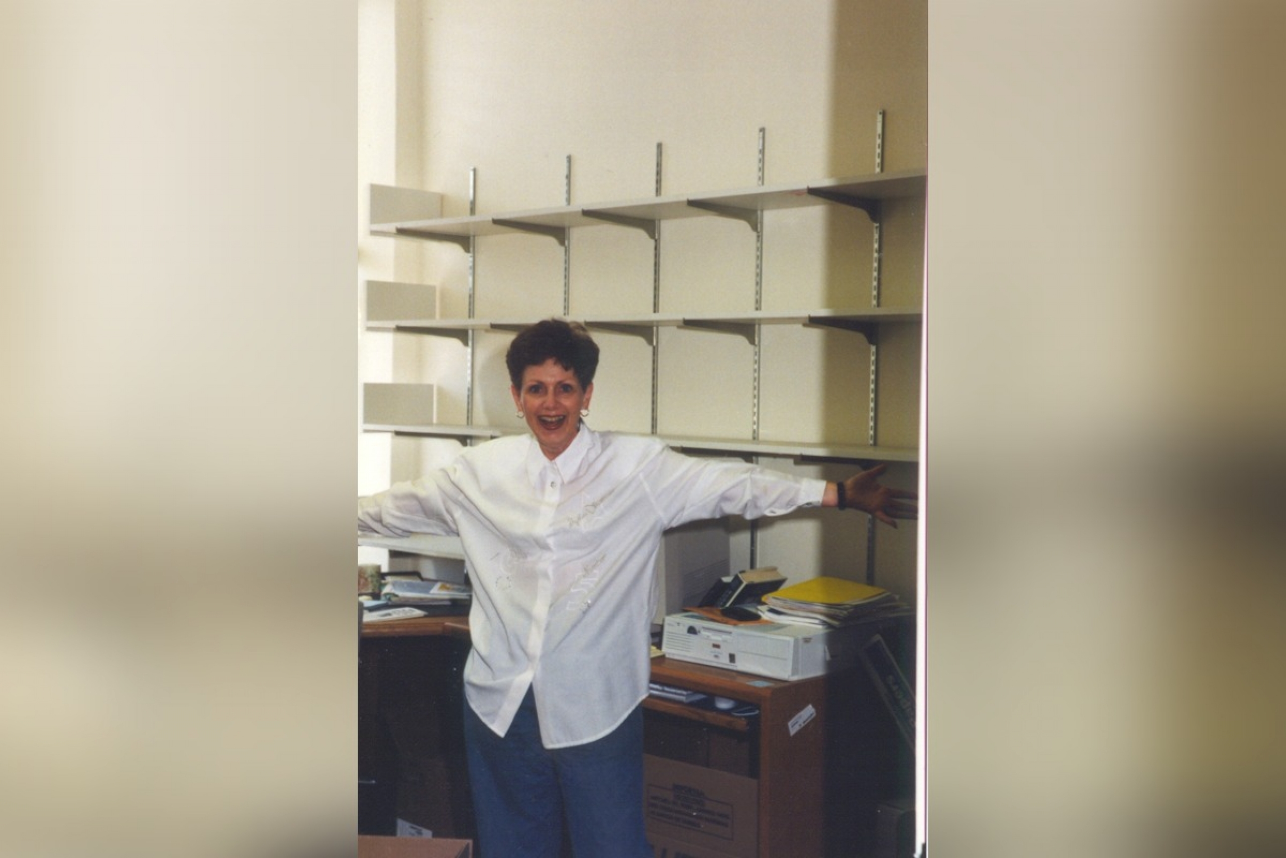 A person smiles at the camera with arms wide open; in the background are empty shelving units.