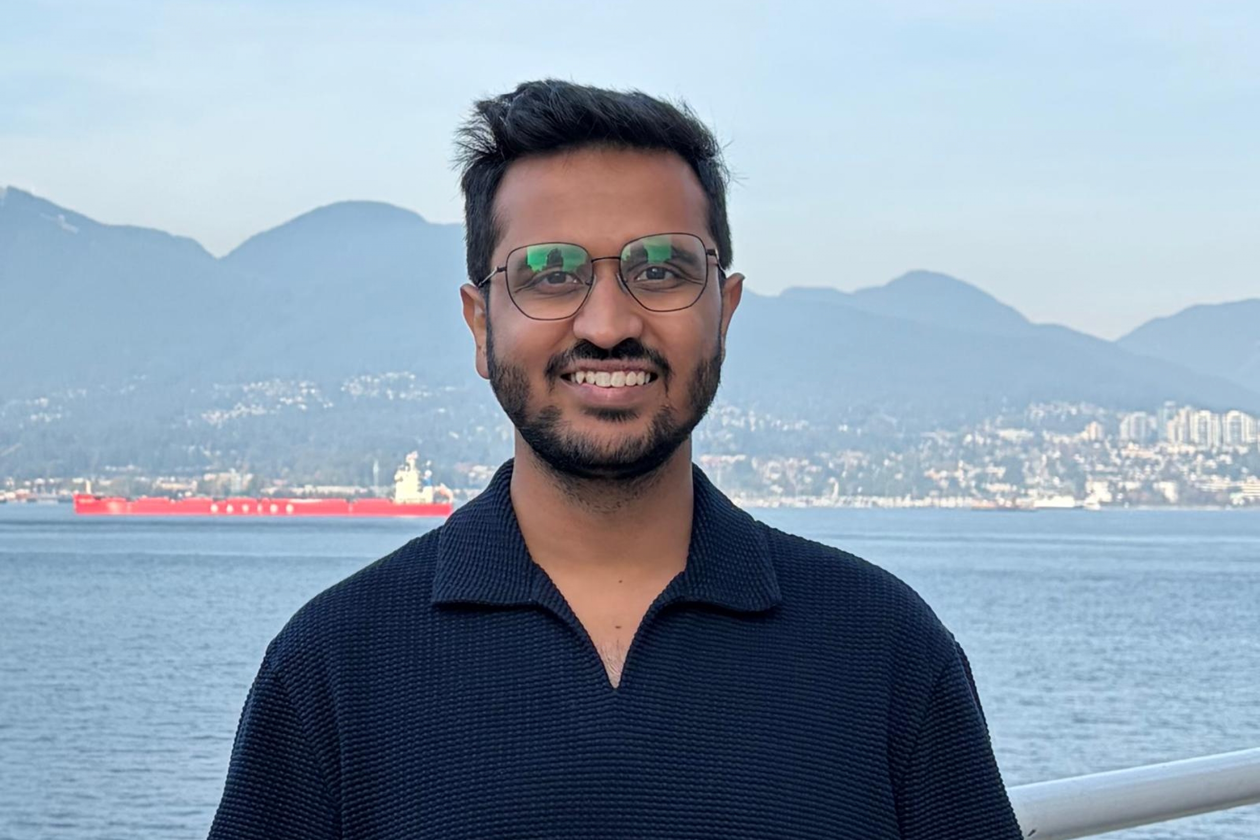 a smiling young man with glasses and a beard stands in front of a harbour front in front of mountains at the foot of which is a city