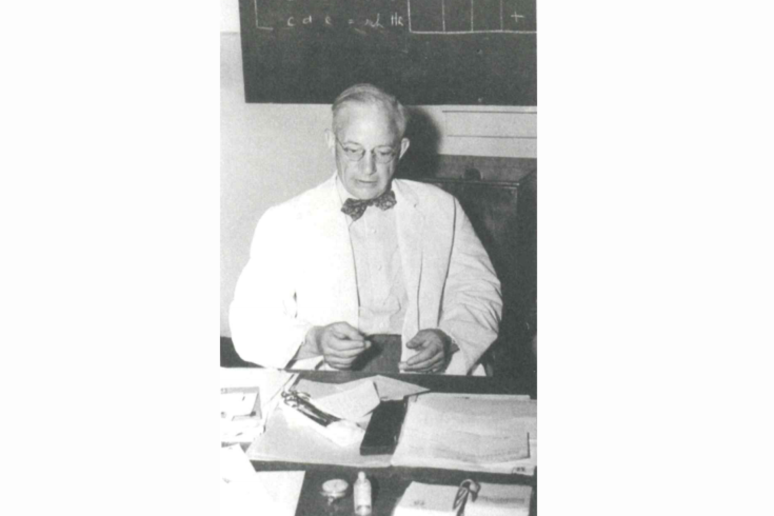 A black and white photo of Dr. Bruce Chown, who wears a white coat and a bow tie in his lab. 