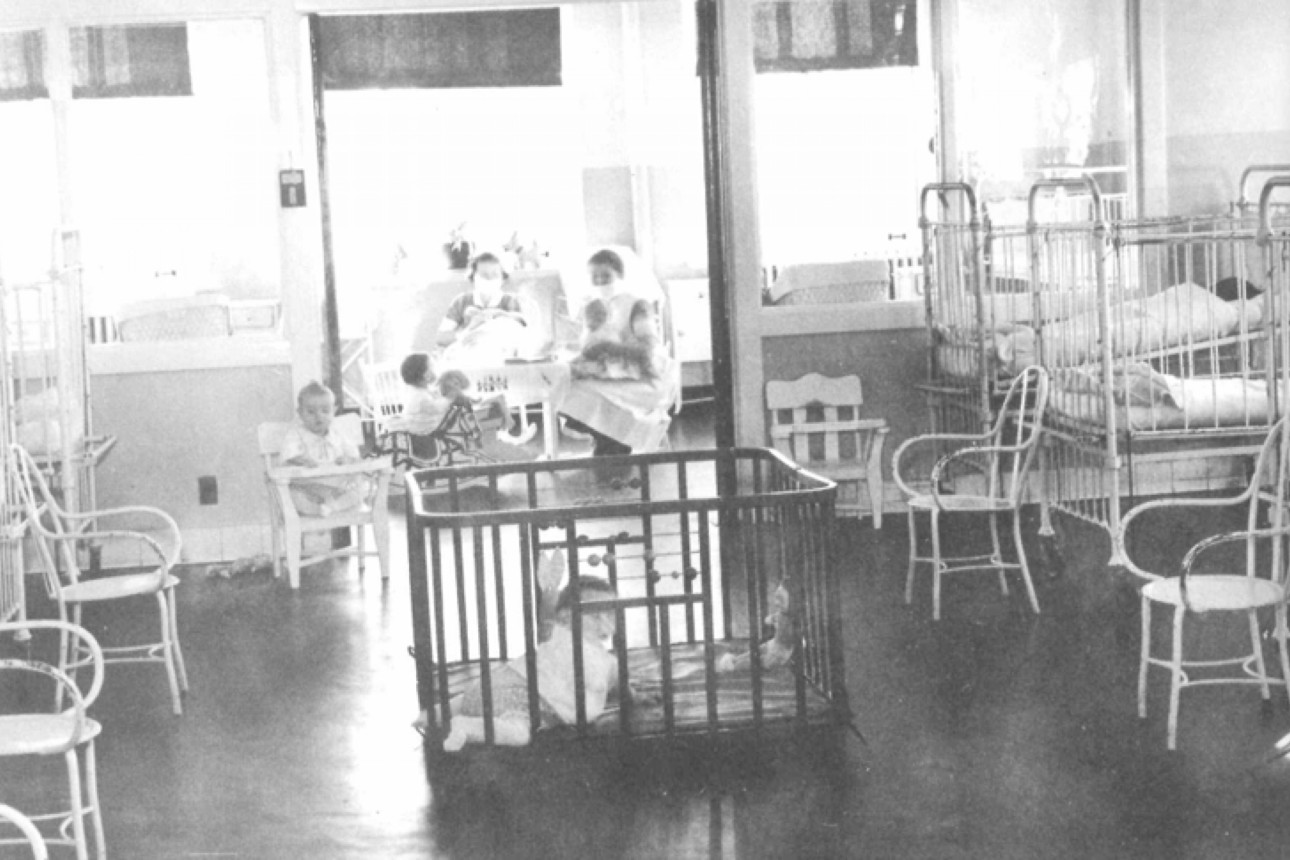 Black and white photo. A child sits in a chair an another one sits in a crib. 