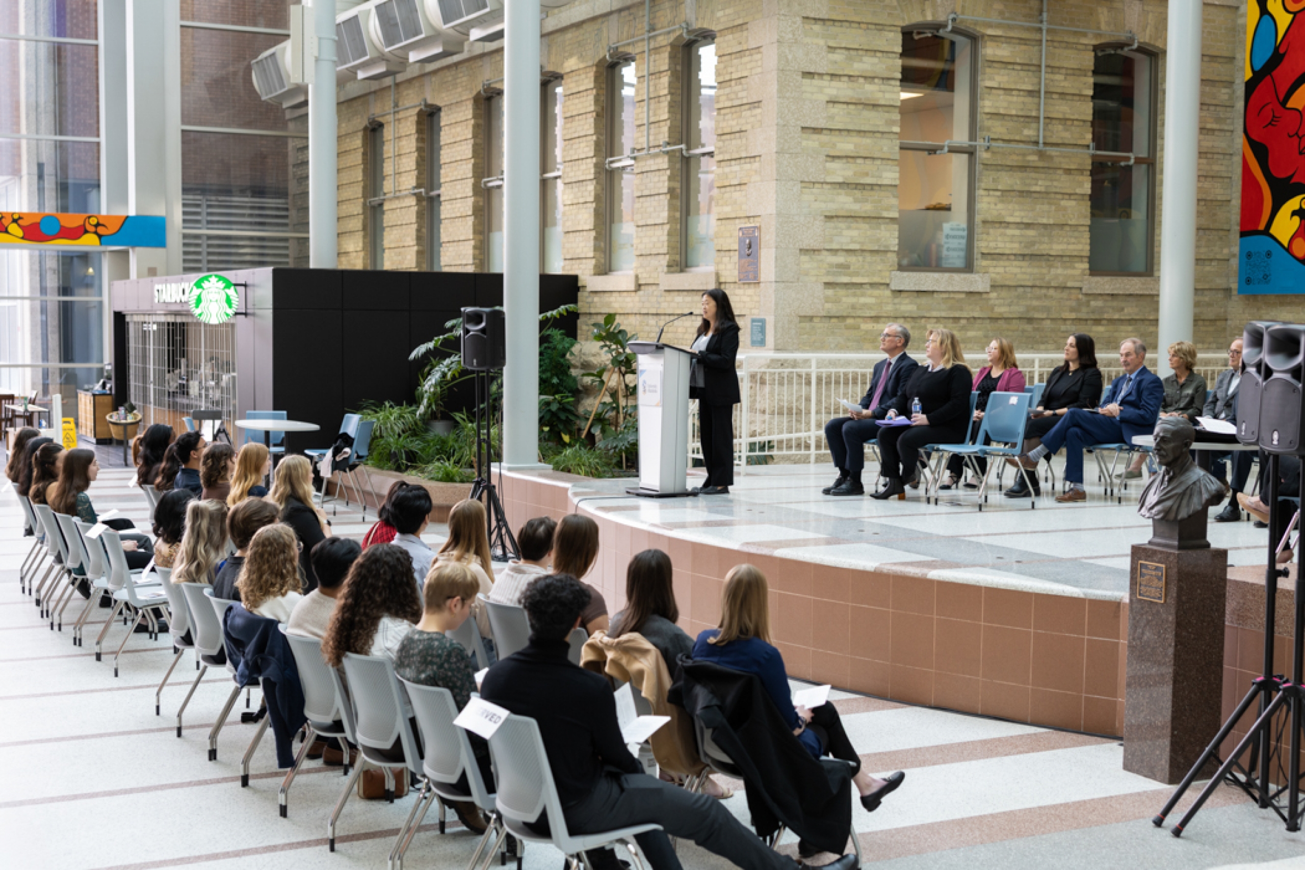 Students sit facing the stage as Dr. Hope Anderson addresses them. Special guests are seated behind Anderson on stage.