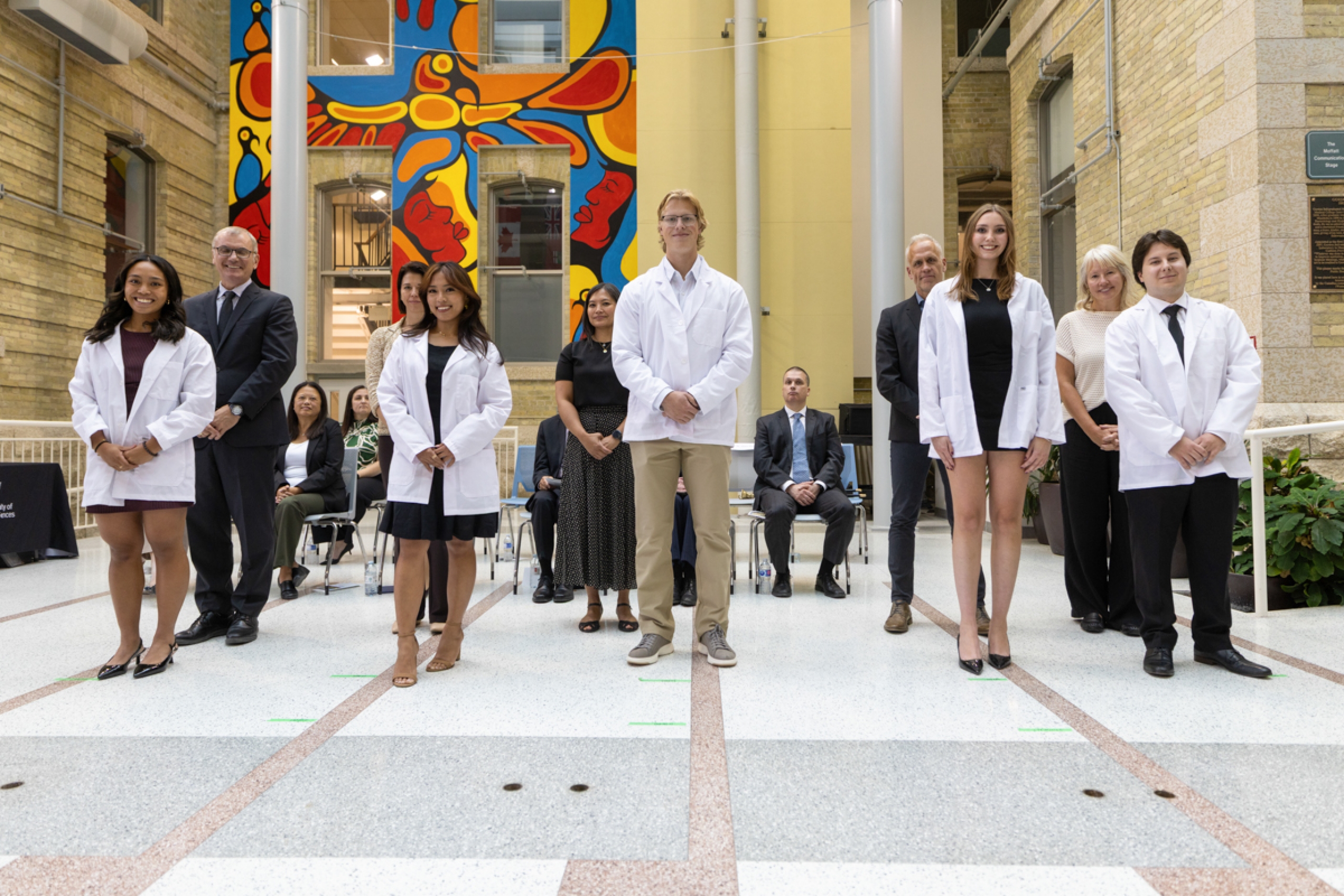 Five students wearing white coats with faculty members behind them.