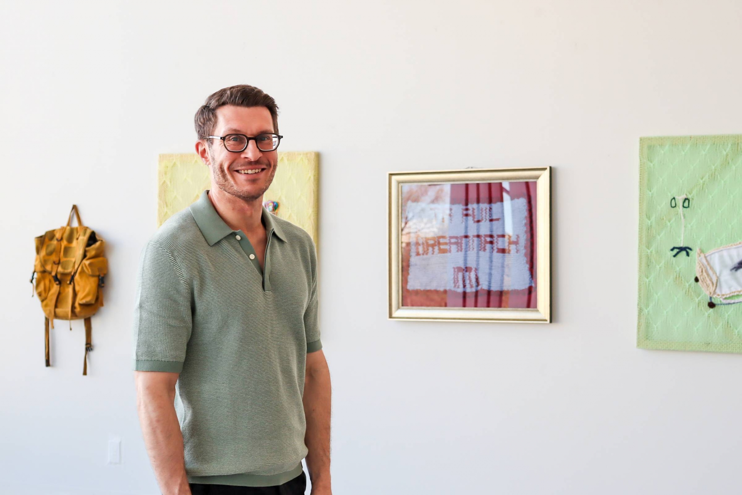 Dr. Stephen Runge stands in the School of Art ARTlab, smiling beside framed artworks installed on a white wall.