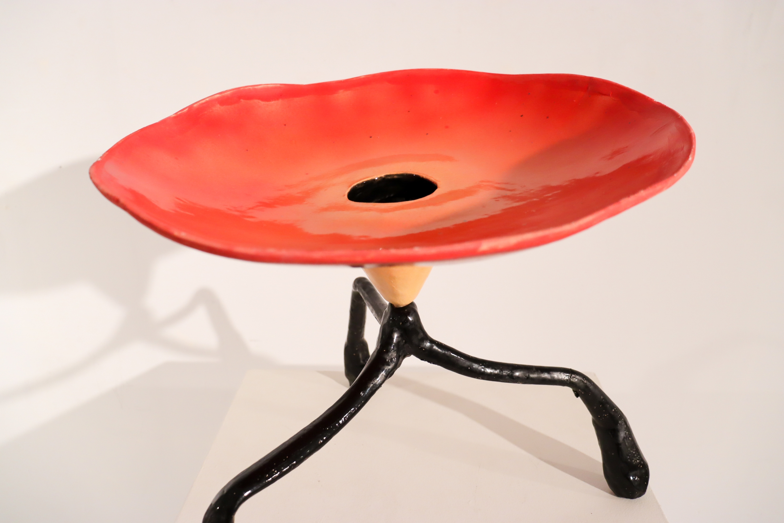 Red ceramic vessel with a wide, shallow bowl and black tripod-like legs displayed on a pedestal in the RECLAYM exhibition.
