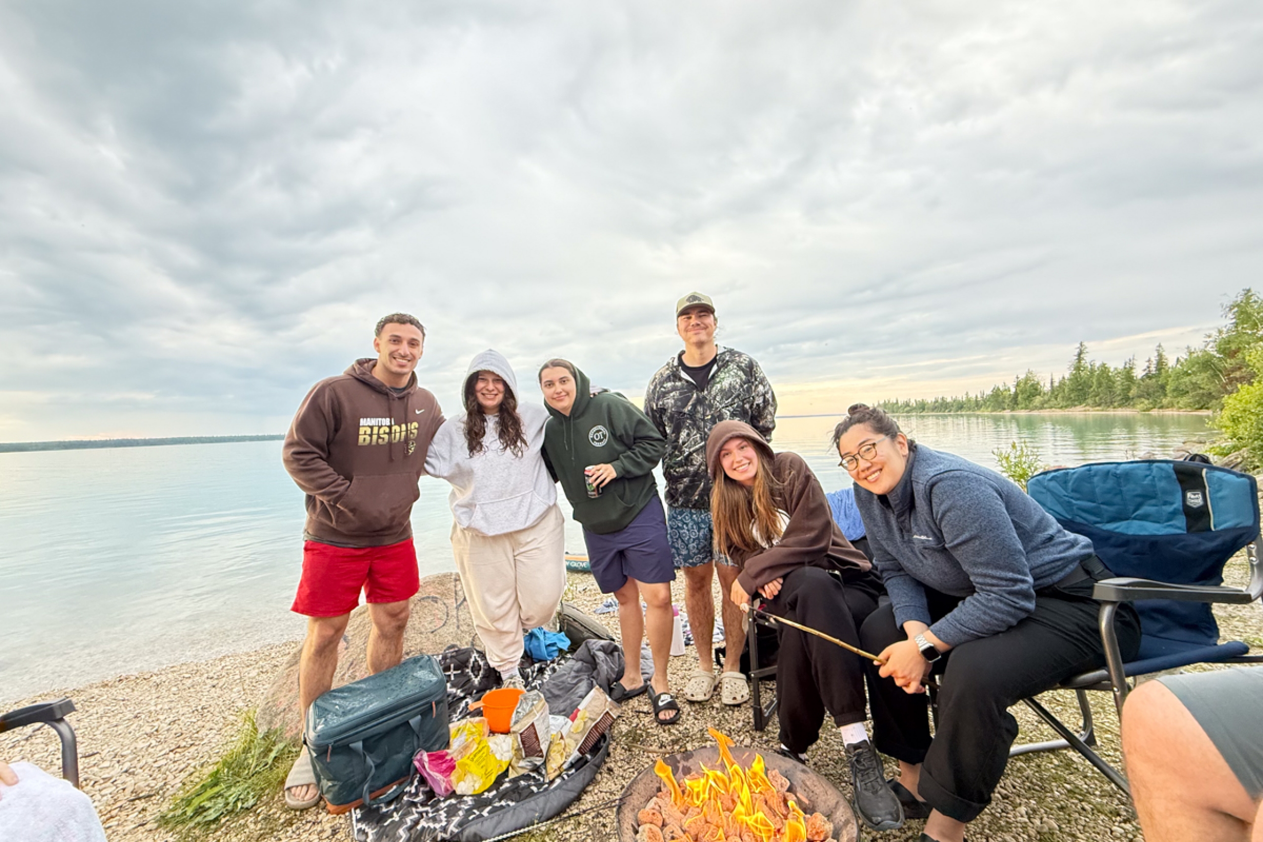 A group of six people gathered around a campfire beside a lake.