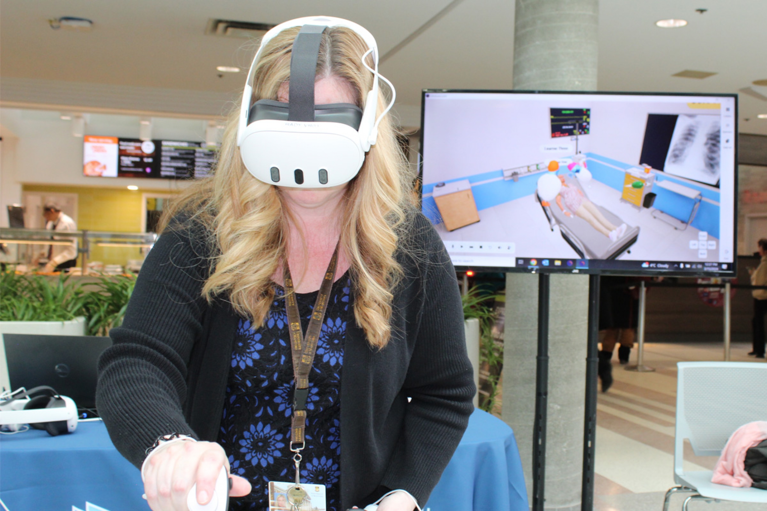 A woman wears a VR headset, while a simulated image of a hospital room displays on a screen behind her.