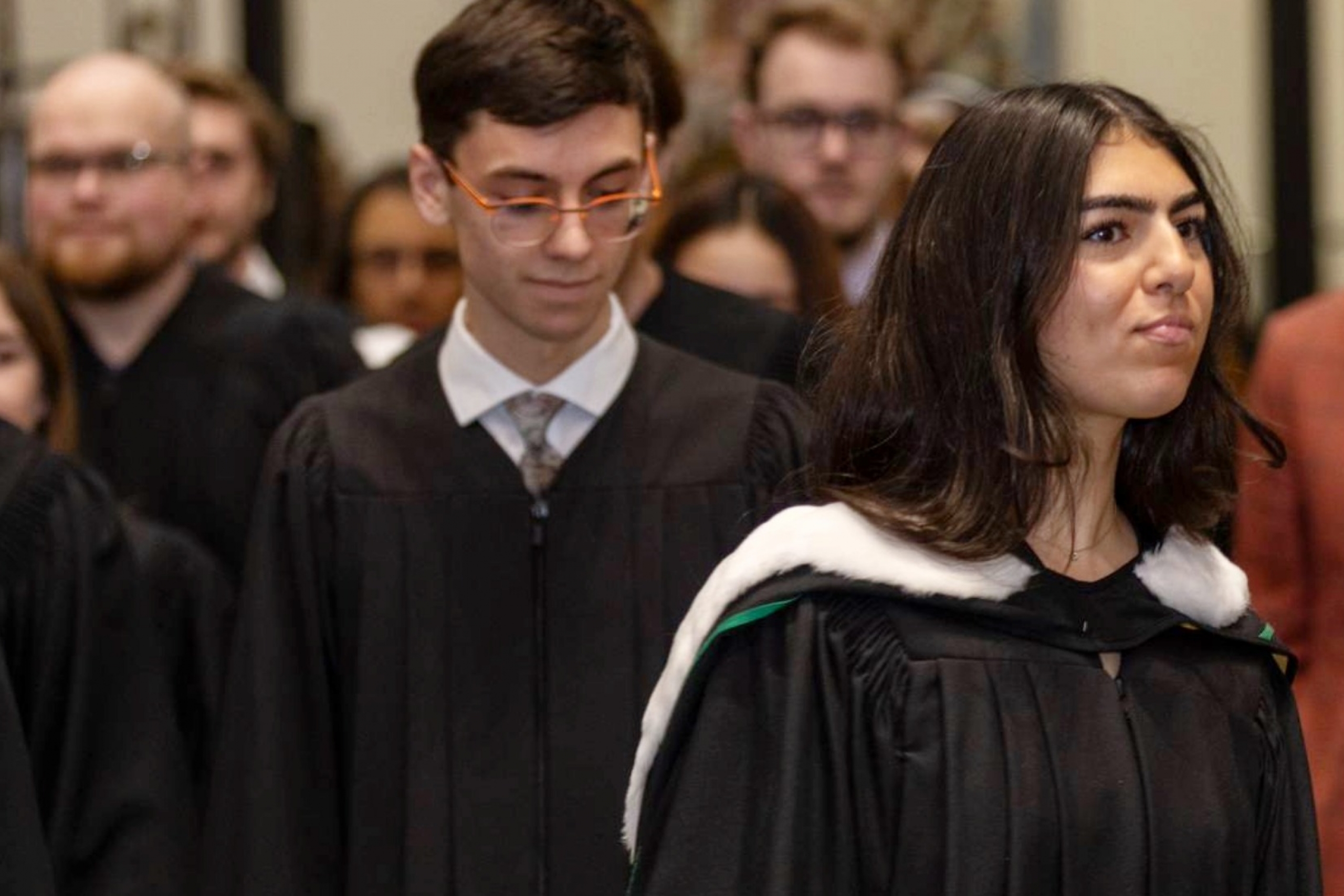Young woman in convocation gown is followed by fellow graduands.