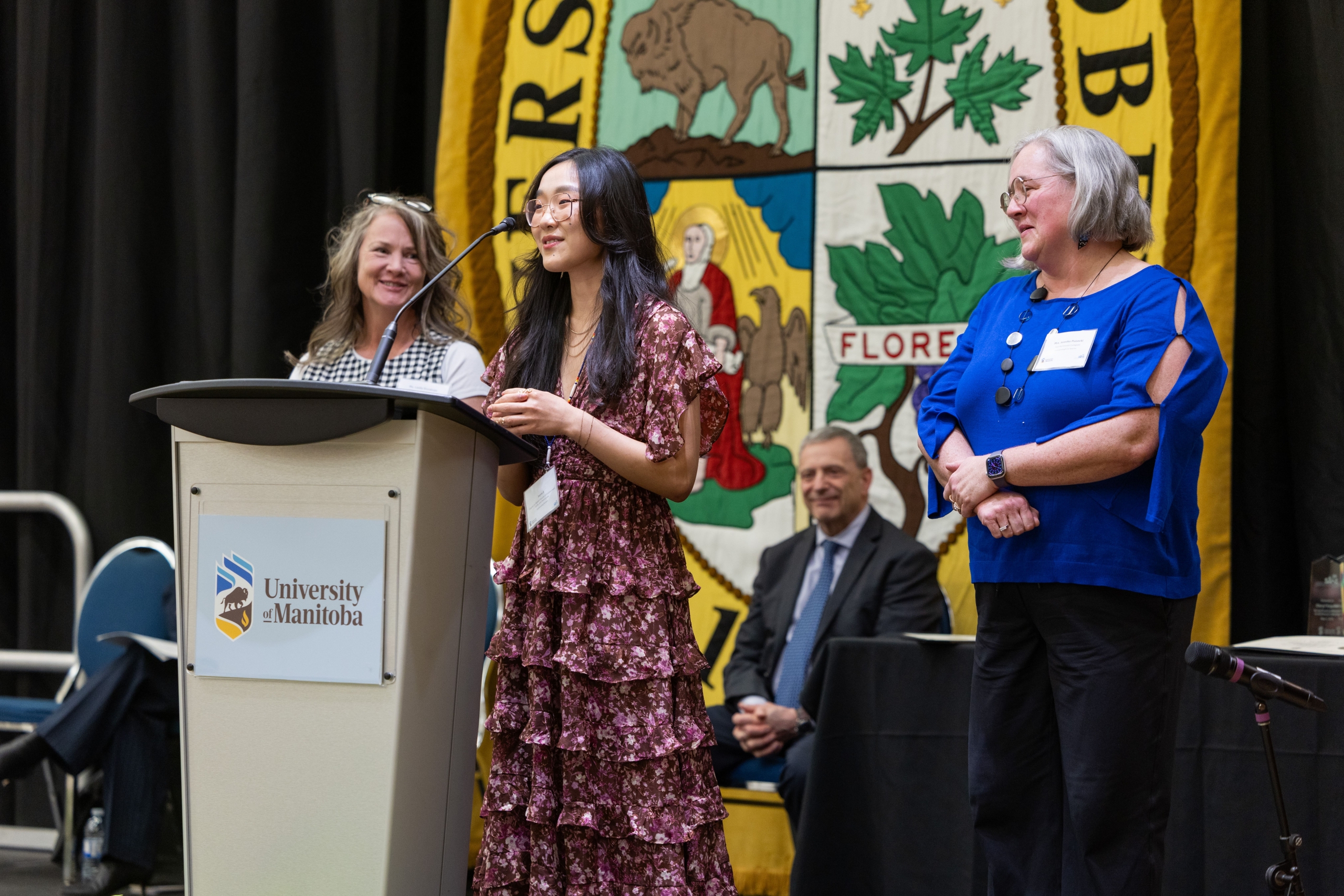 A graduating student stands before a podium to give a speech next to her former educators.