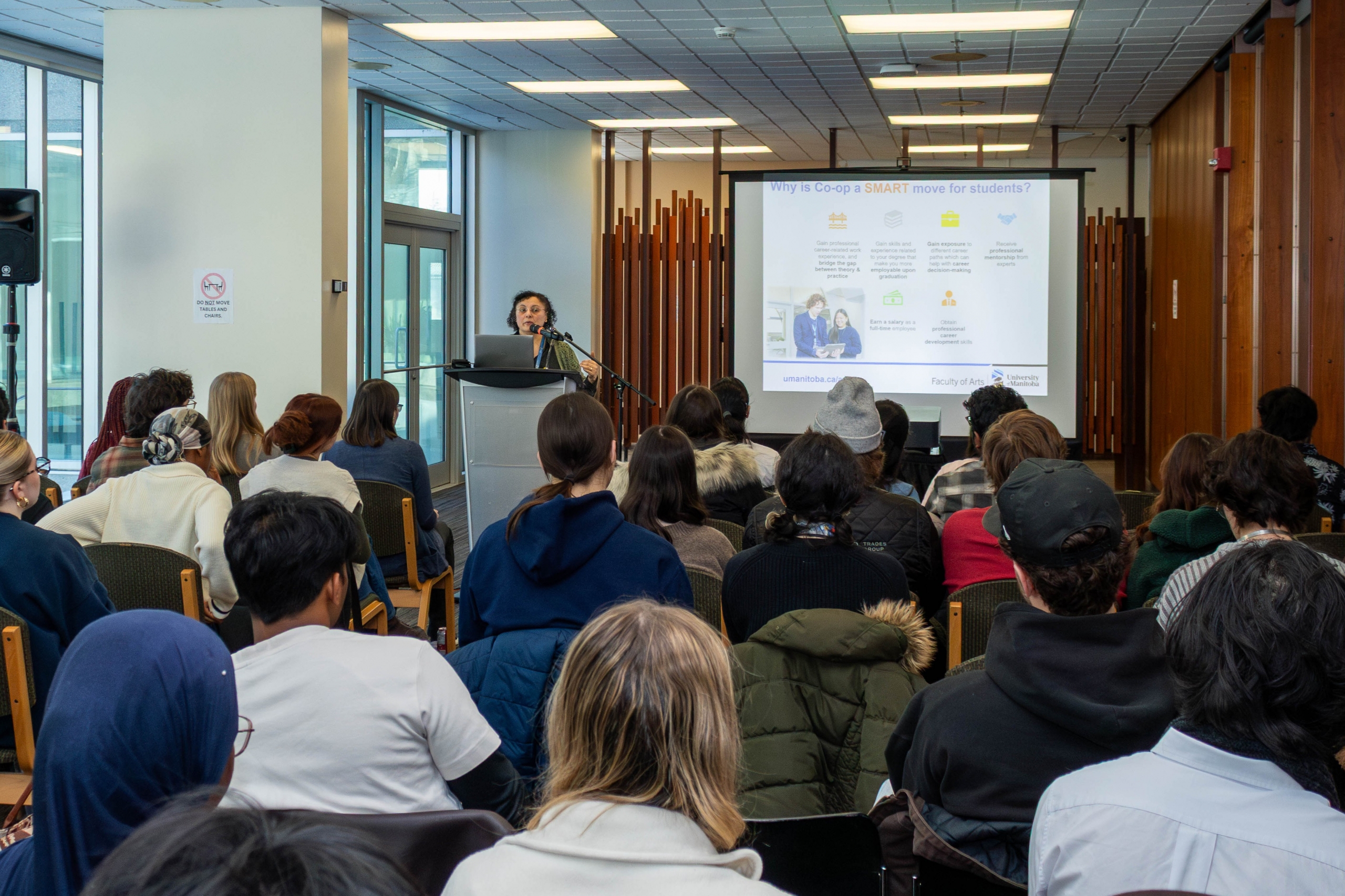 A room full of students watching a presenter at a podium with a screen beside her.