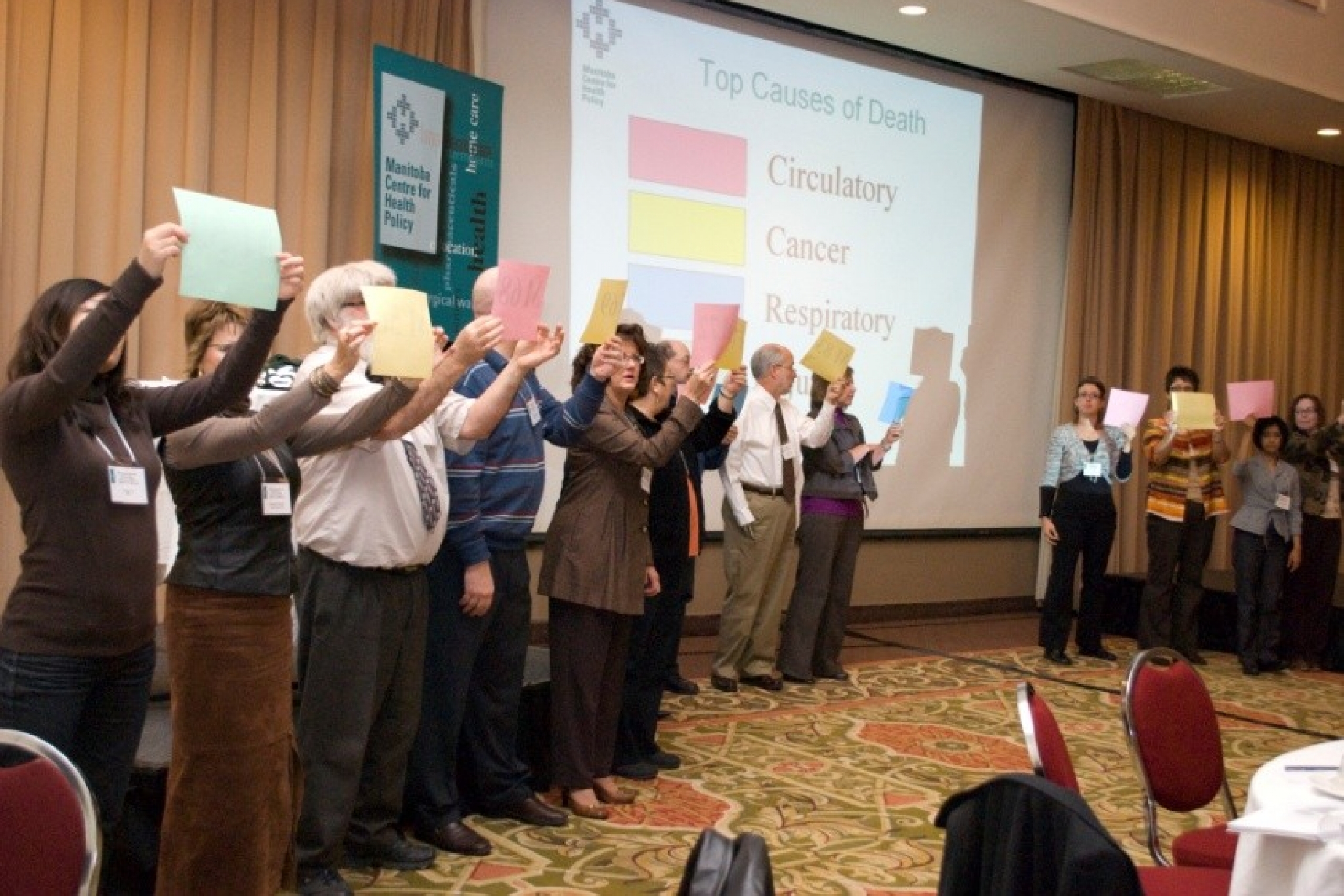 A group of people in front of a room holding up paper. Displayed behind them onscreen is: "Top Causes of Death: Circulatory, Cancer, Respiratory". 