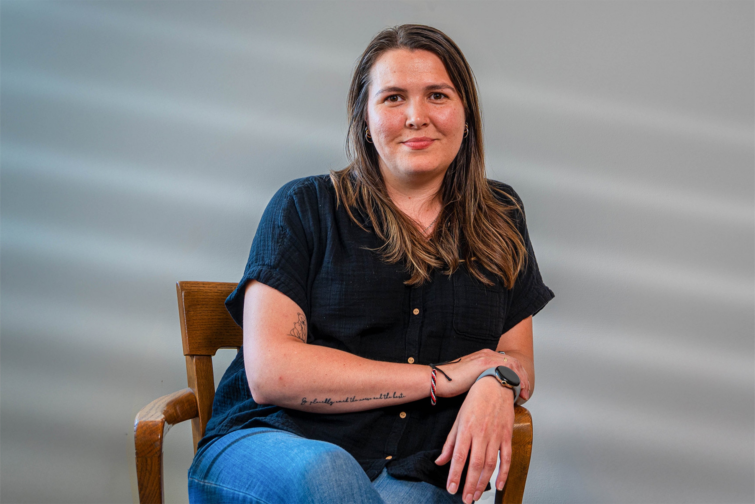 Woman sitting on wooden desk chair, one arm resting on top of the other, smiling at the camera.