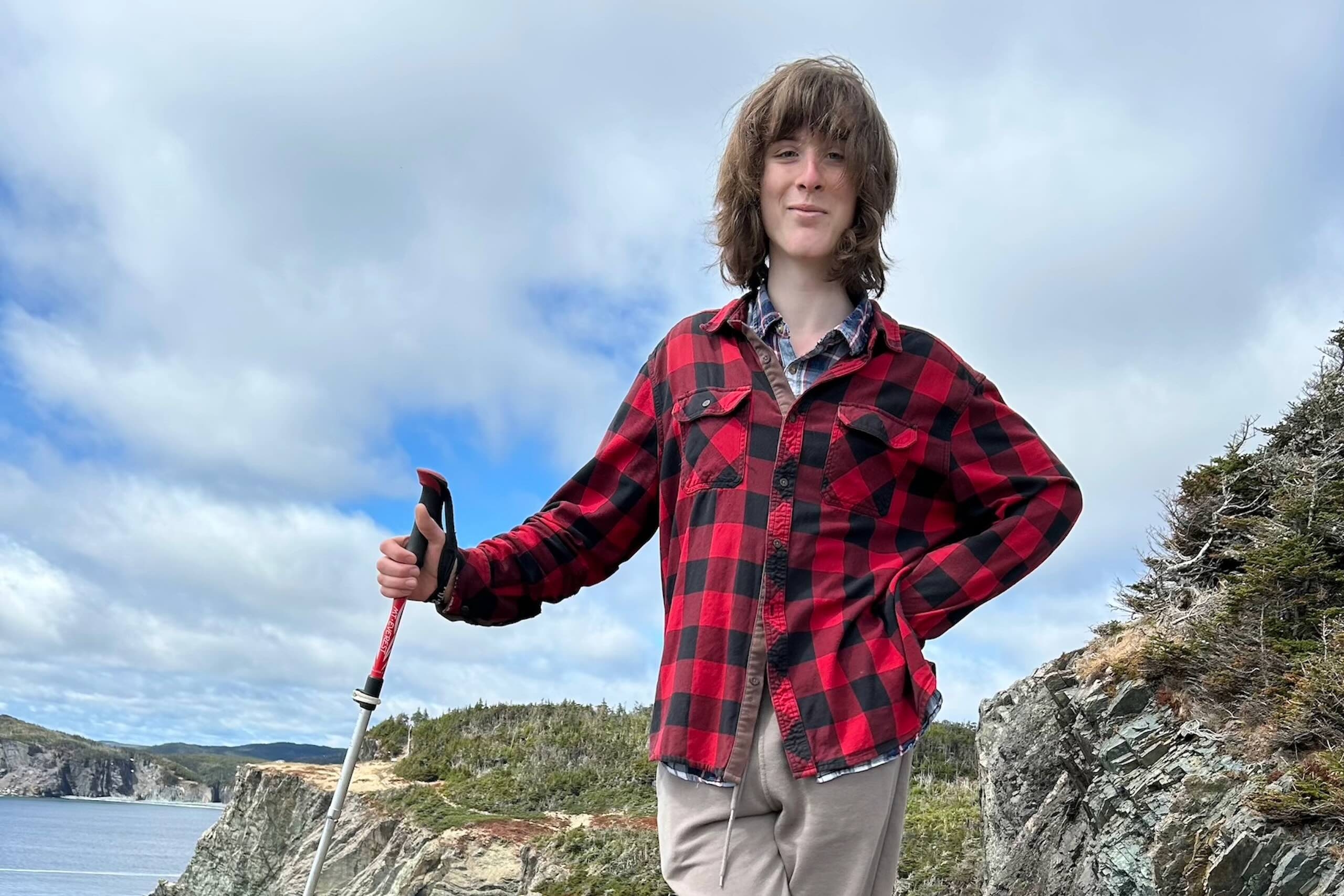 Smiling young person in red buffalo check jacket standing atop a craggy cliff with sky and sea in background.