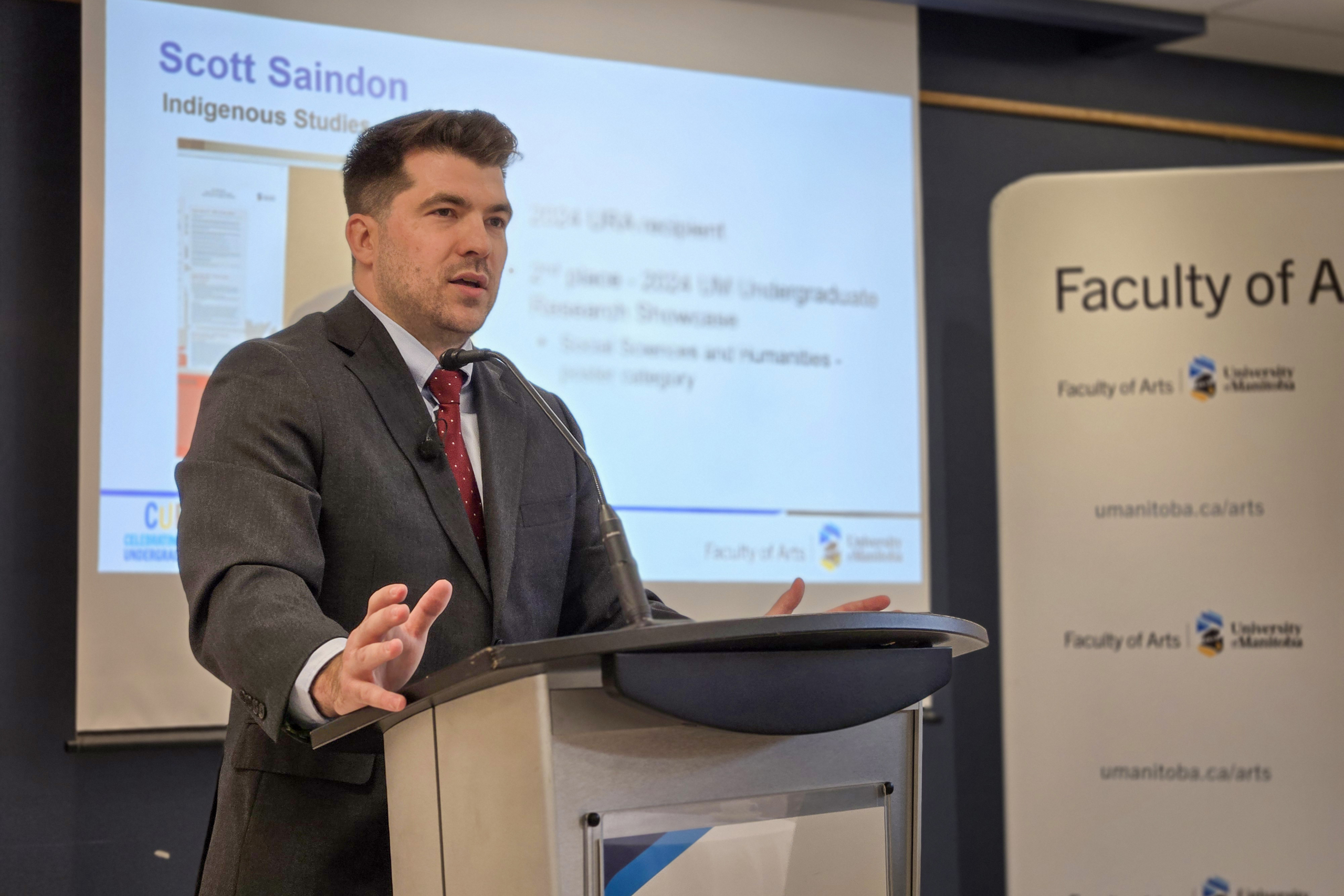 Male wearing a suit standing behind a podium, speaking to the room.