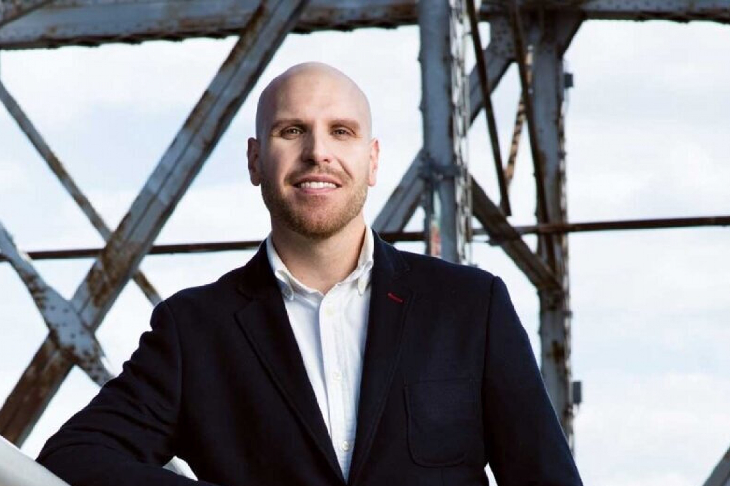 Bald man with a short beard, wearing black suite, white shirt leaning on a guard rail.