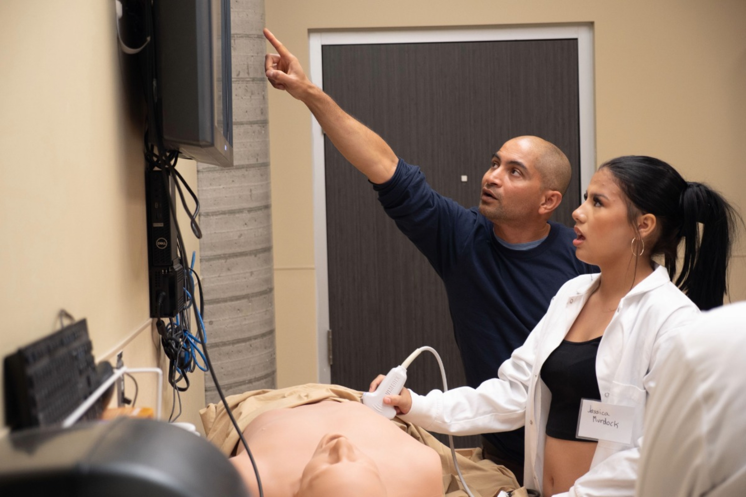 A learner uses an ultrasound probe on a manikin as an educator points to the ultrasound monitor.
