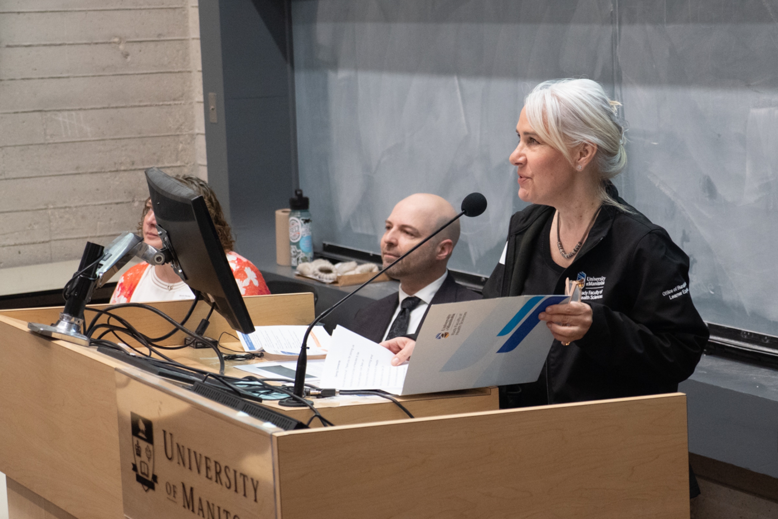 A person talking in front of a podium with two people seated beside.