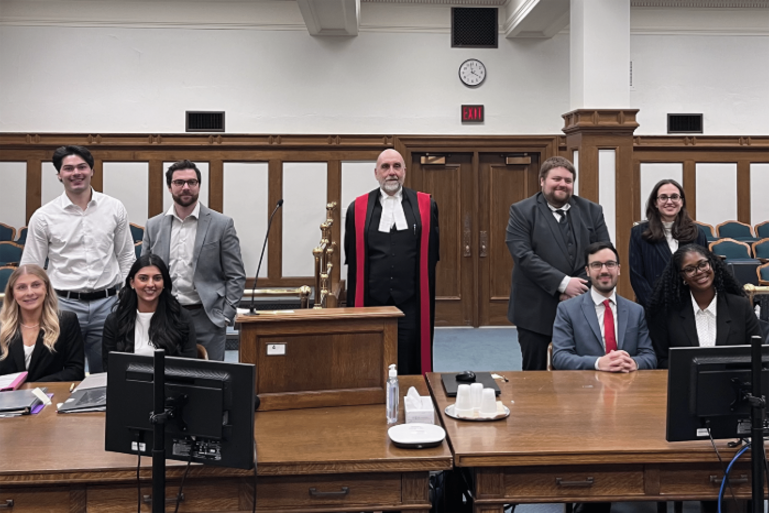 a group of students sit and stand in a courtroom on each side of a judge in robes who stands in the middle.