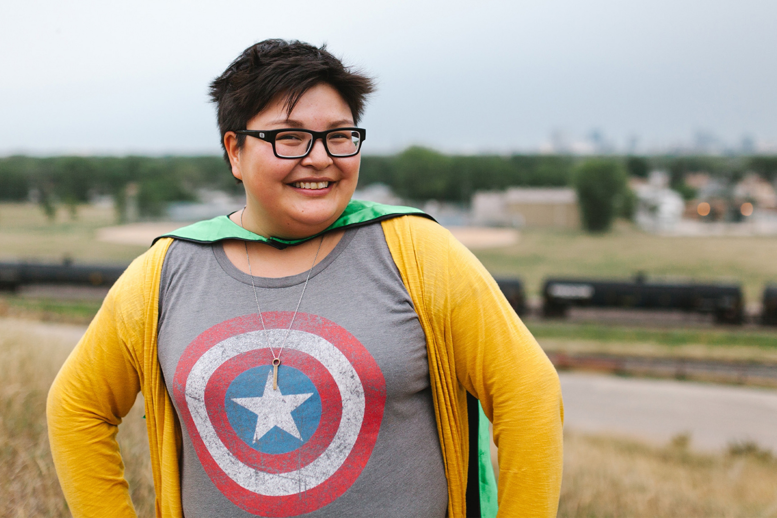 Woman wearing glasses, a tshirt with the Captain America shield on it and a cape, stands with her hands on her hips.