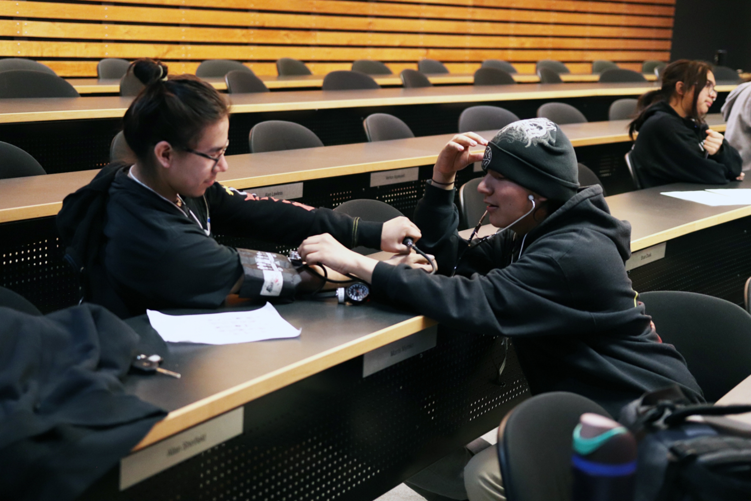 The person on the right is checking the other person’s blood pressure and listening to their vitals with a stethoscope.