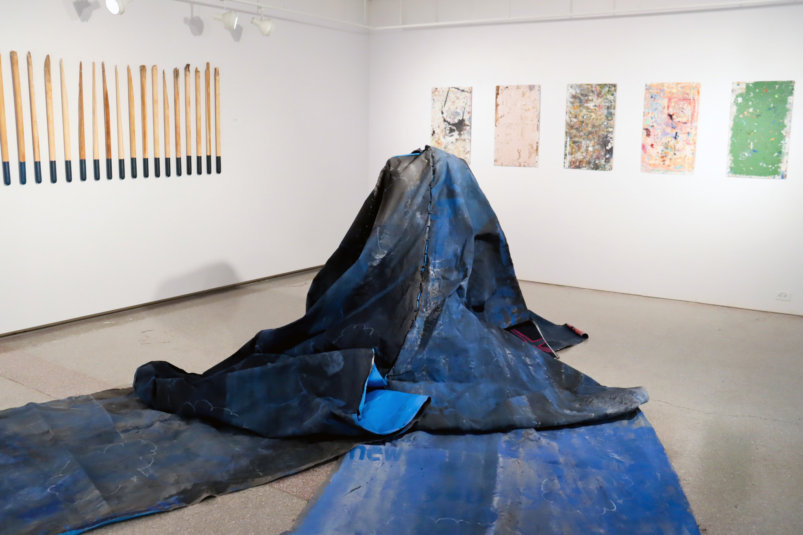 Installation view with large blue tarp-like form on the floor and wall-mounted works.