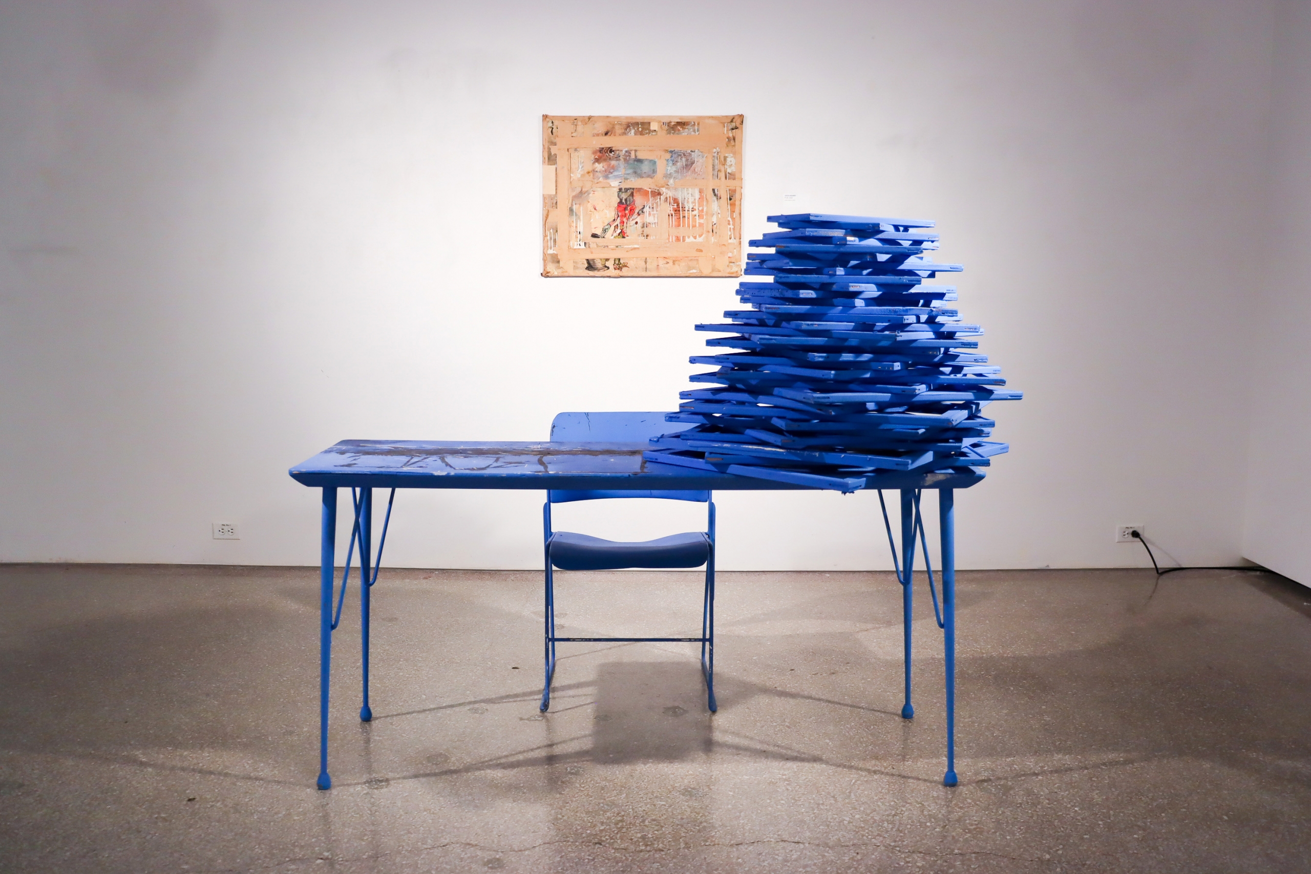 Installation view featuring a blue table and stacked painted panels with a framed painting on the wall behind.