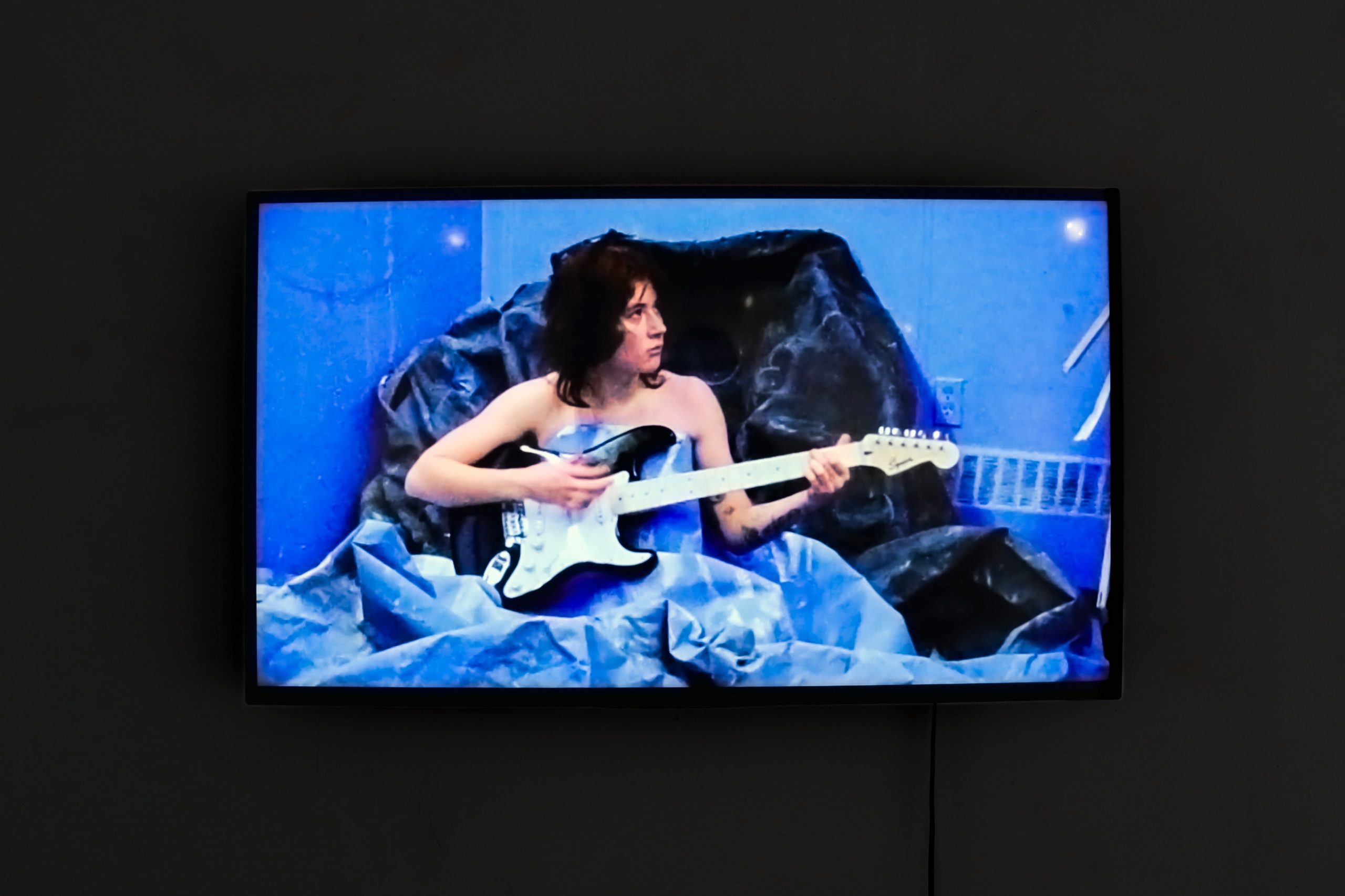 Video installation showing a figure seated with an electric guitar in a blue-toned environment.