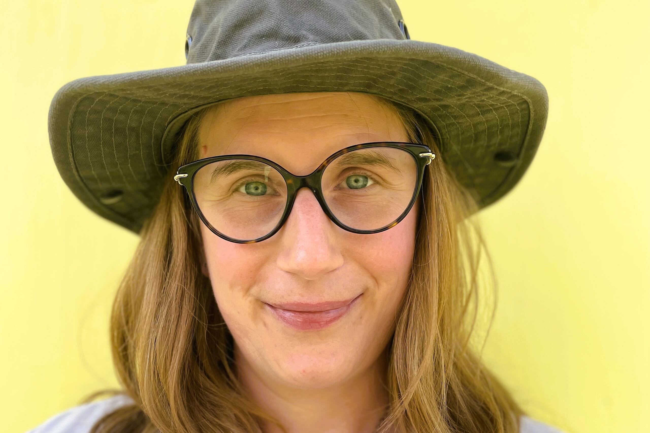 Smiling person with glasses and long hair wearing a field hat.