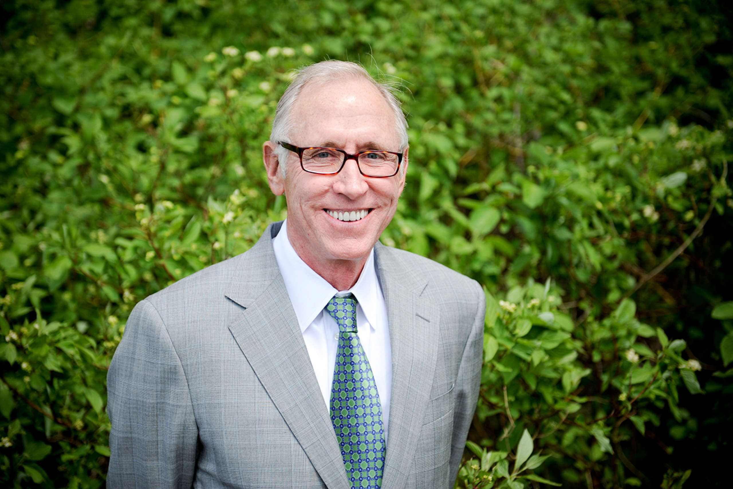 An older man with glasses poses in front of greenery