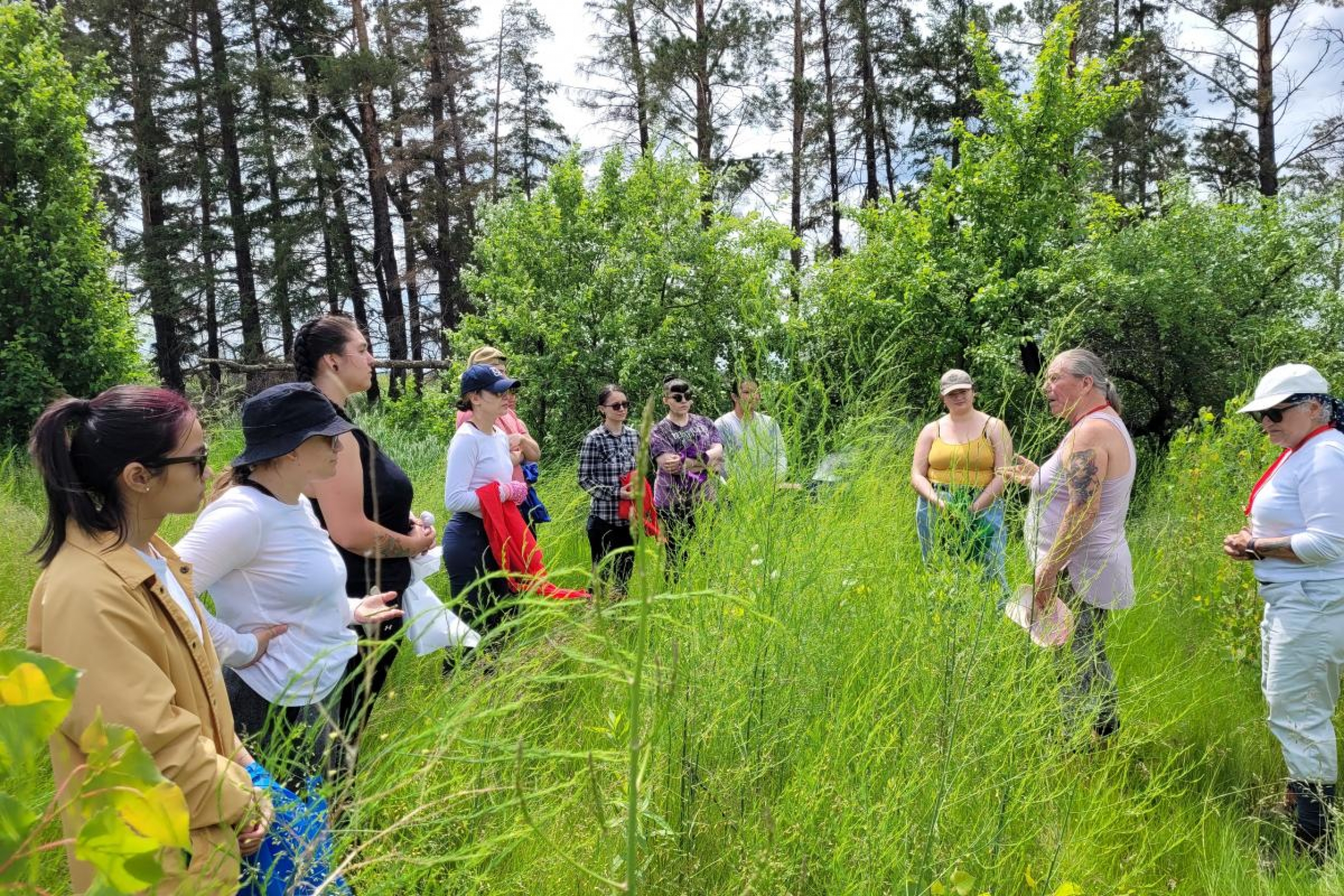 students standing in tall grass, listening to Elder speak.