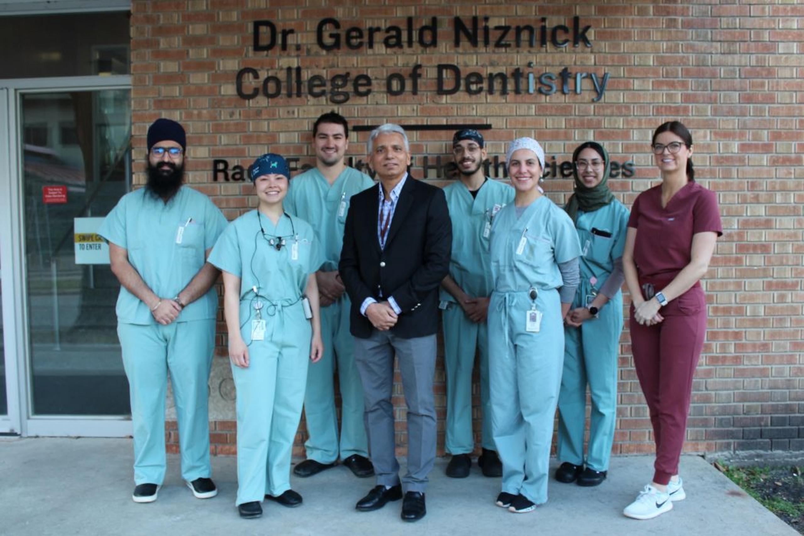 Eight people stand outside. Seven of them are wearing scrubs and one is wearing a suit. On a wall behind them reads "Dr. Gerald Niznick College of Dentistry."